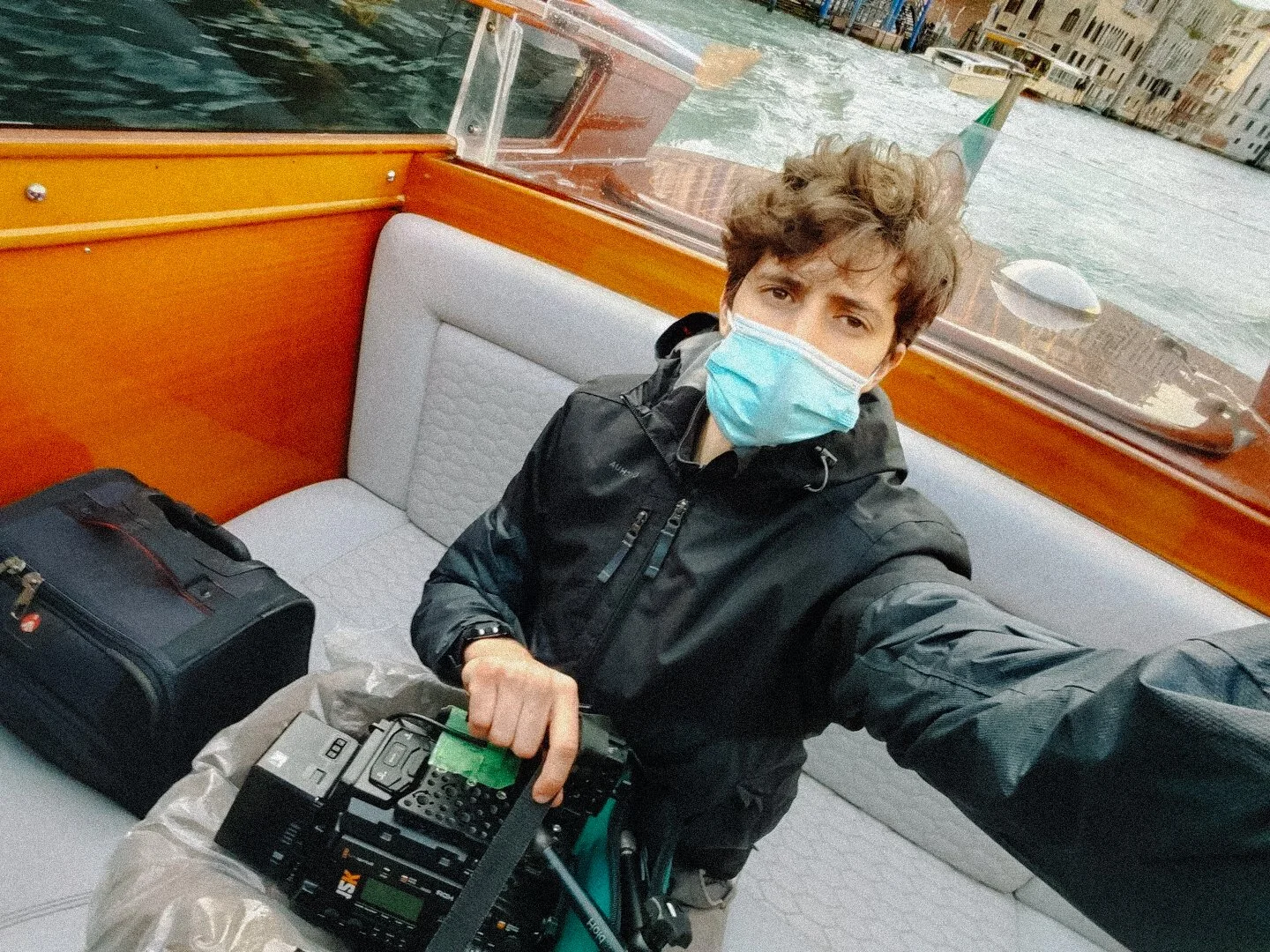 Person wearing a mask on a boat holding a camera, bag beside them, Venice canal in background.