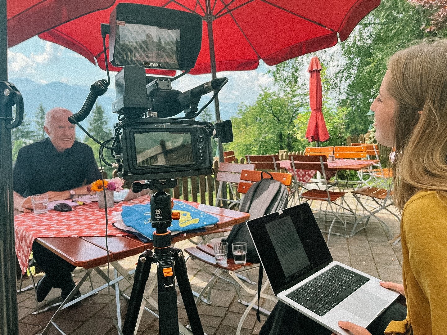 Outdoor interview setup with a man sitting at a table under a red umbrella, a camera on tripod facing him, and a woman with a laptop taking notes.