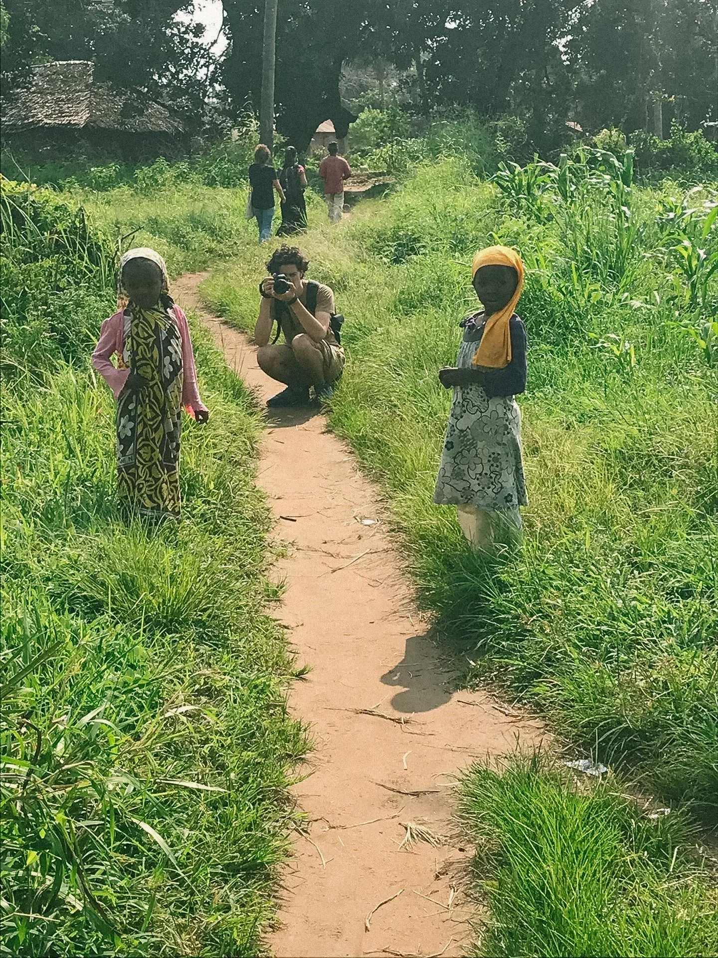 A photographer kneels on a dirt path capturing two children in a grassy rural area.