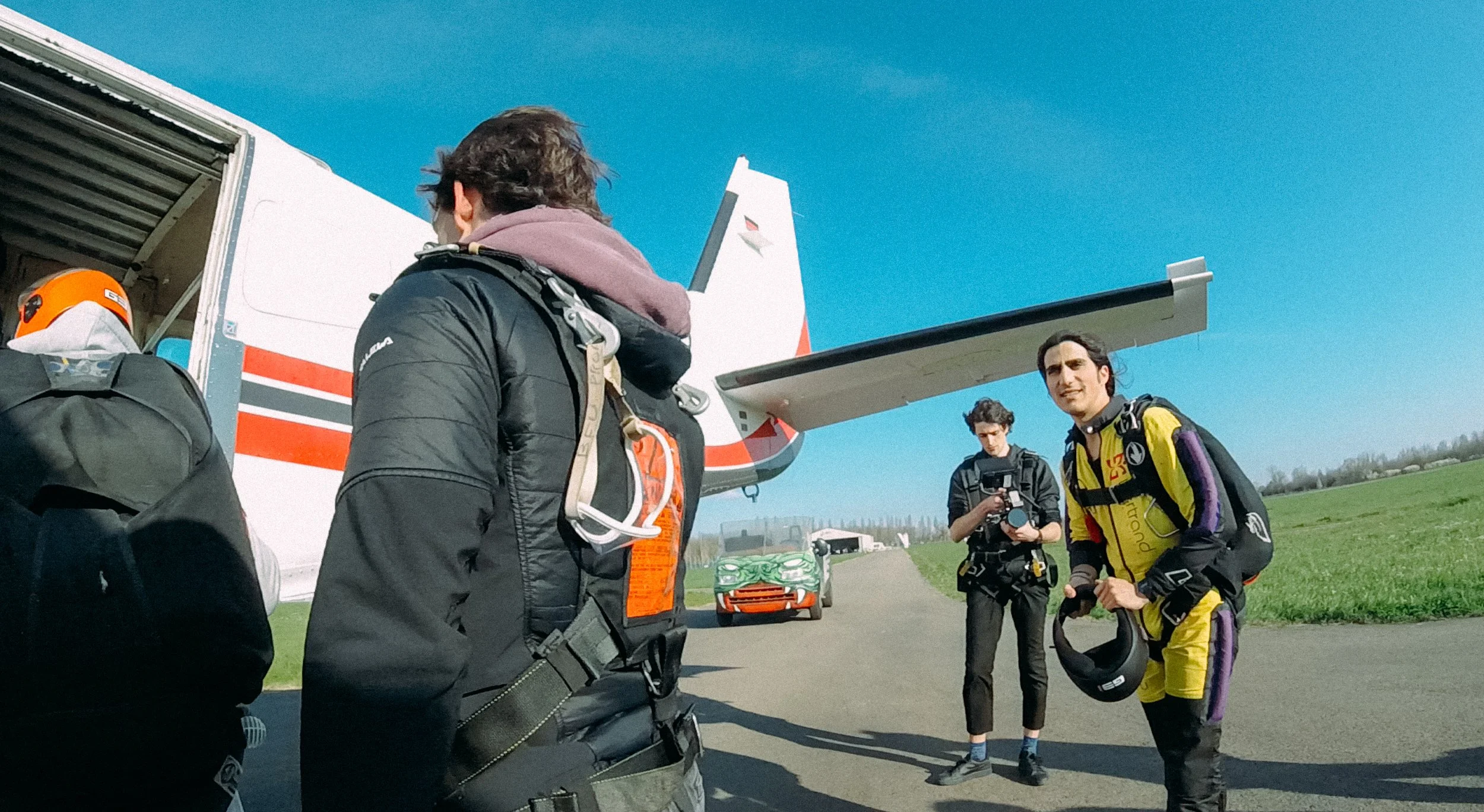 Skydivers preparing to board a plane, wearing jumpsuits and parachutes, at an airfield on a clear day.