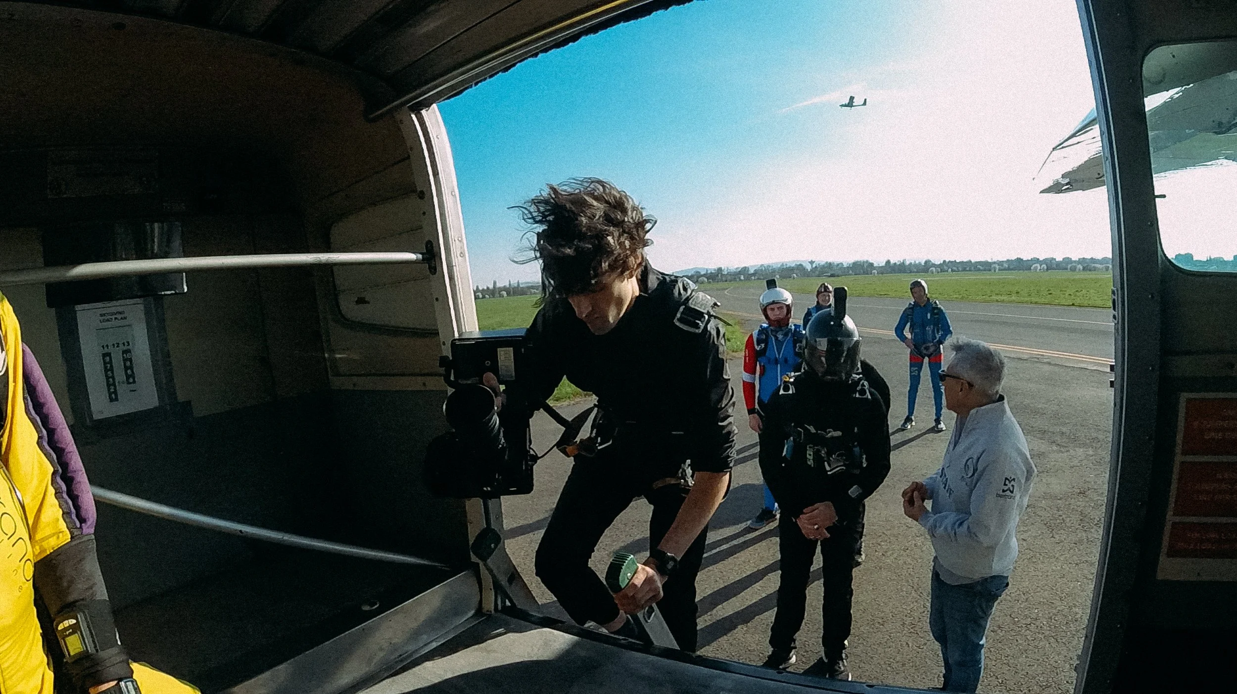 People boarding an aircraft for skydiving, with a cameraman entering, others in jumpsuits waiting outside, and a plane in the sky.