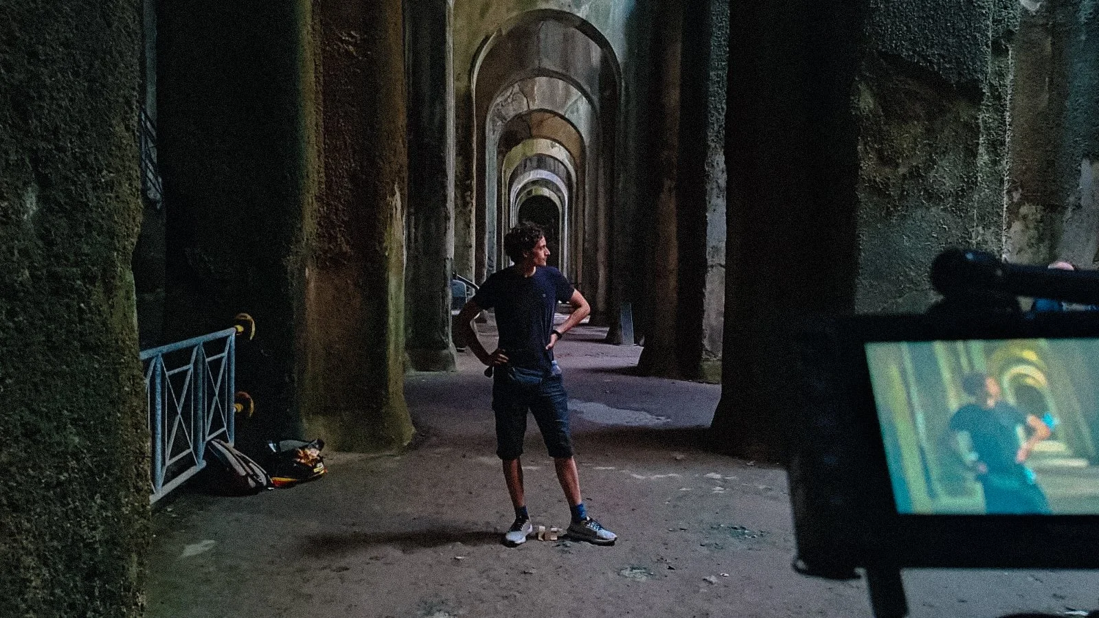 Person standing in a dimly lit, vaulted passageway with stone arches, captured on a camera's viewfinder. The scene includes moss-covered walls and a bag on the ground.