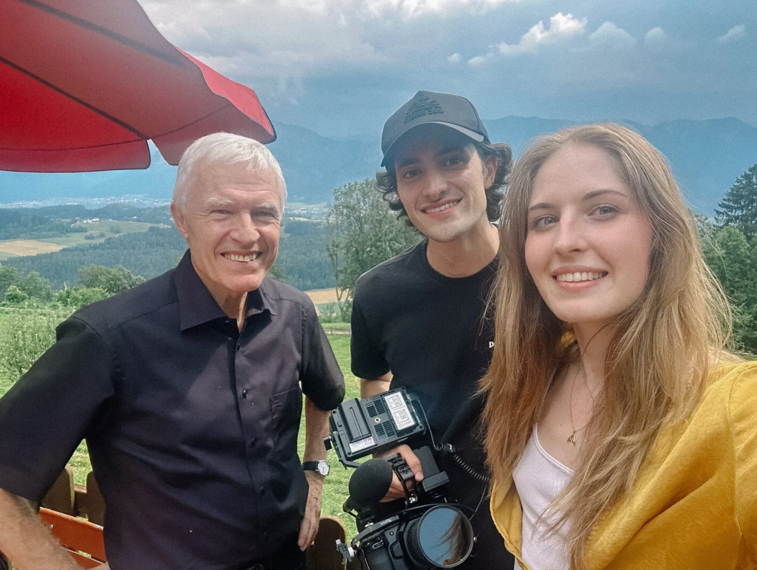 Group selfie outdoors with mountains and greenery in the background, featuring three smiling people; one holding a video camera.
