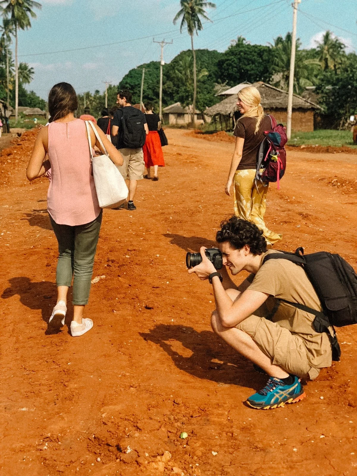 Group of people walking on a red dirt road, person crouching with a camera, tropical scenery in background.