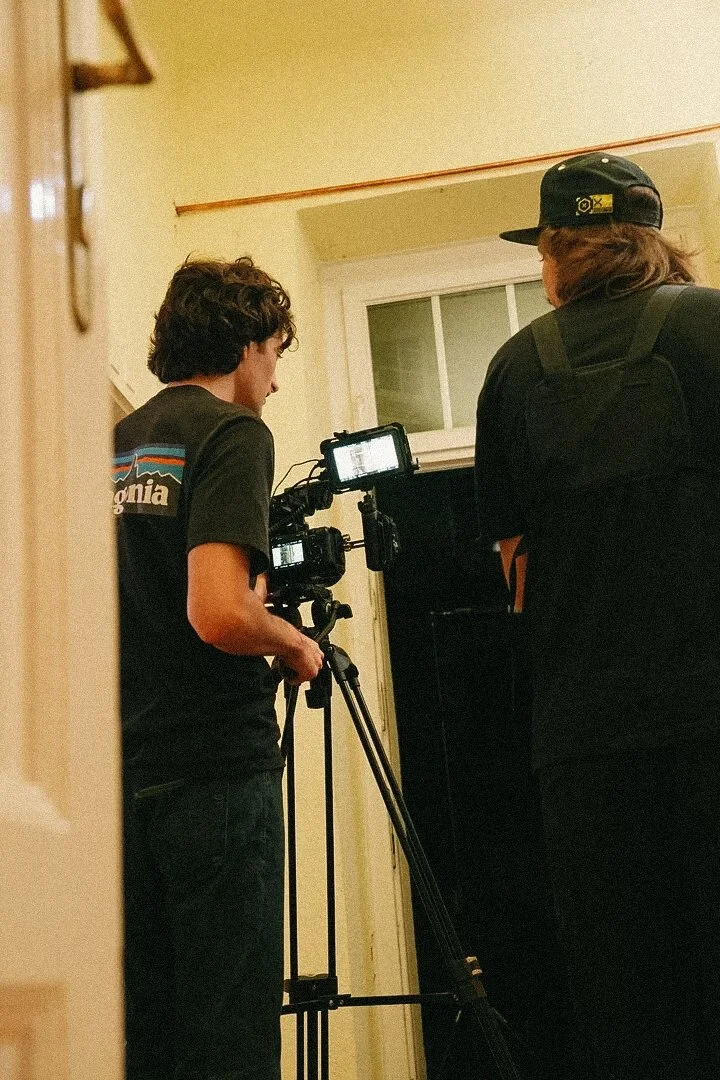 Two people operating a camera on a tripod in a room with a window.