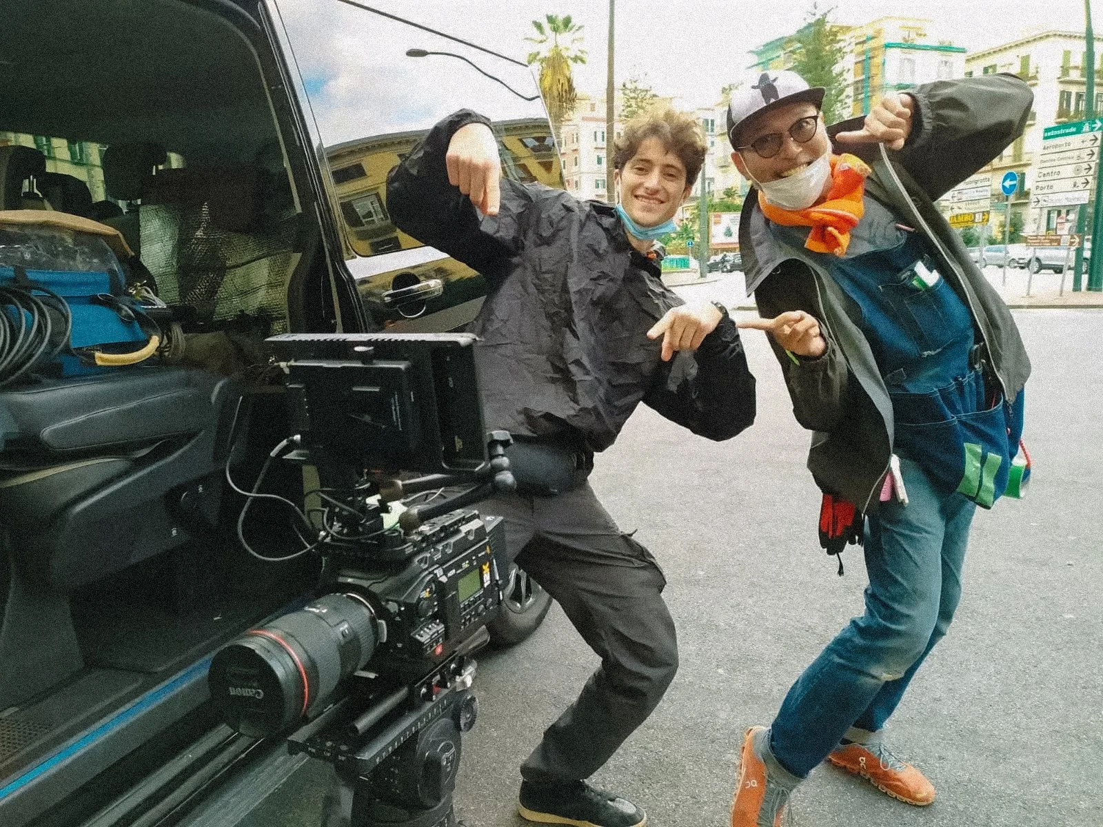 Two people posing next to a professional camera setup mounted on a tripod outside a vehicle, with buildings in the background. They are making playful gestures and one is wearing a face mask on their chin.