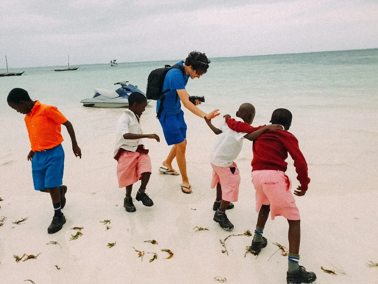 Group of children and an adult playing on a sandy beach by the ocean with a jet ski in the background.