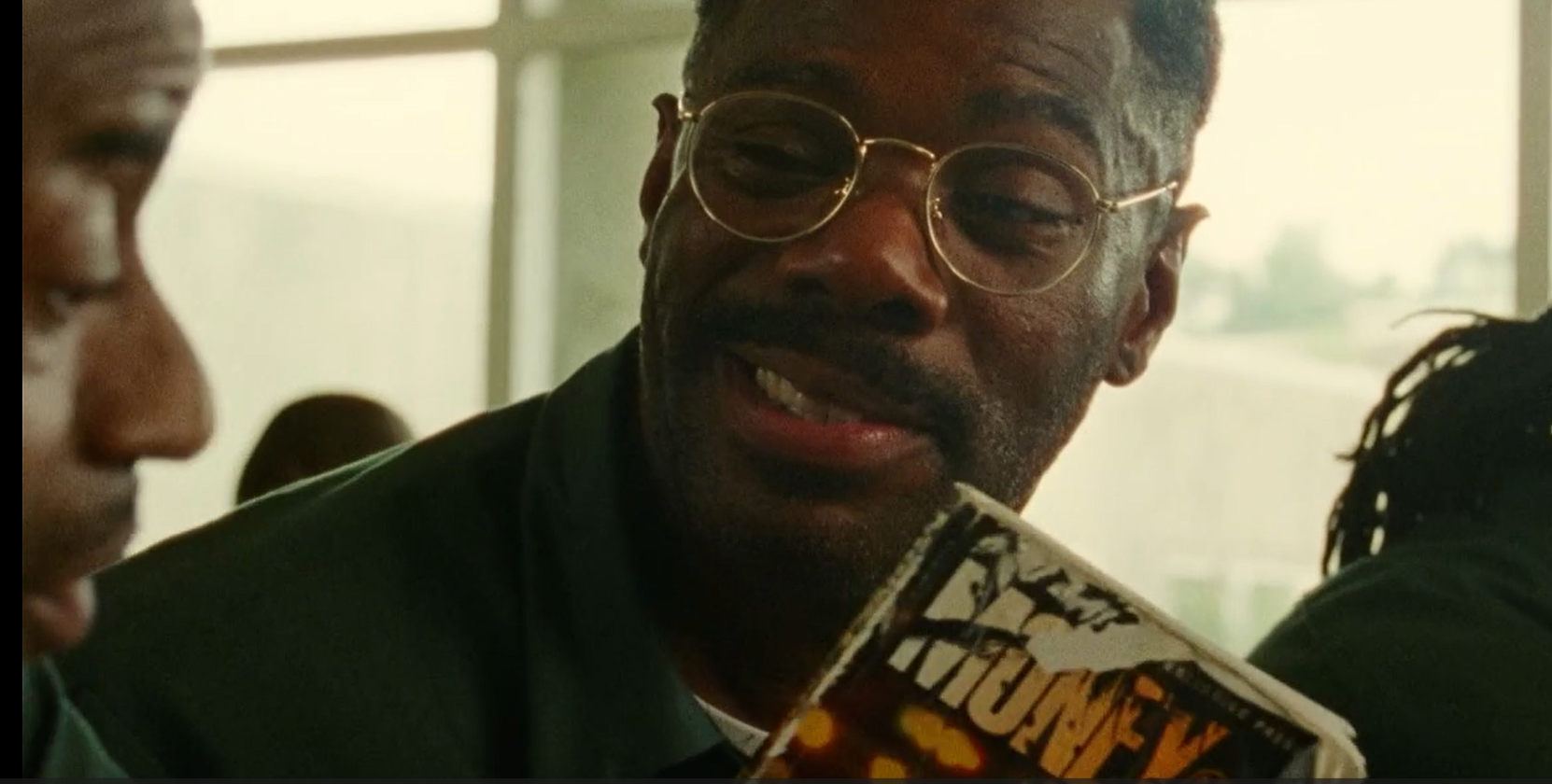 Man wearing glasses, smiling, holding a wore novel, sitting indoors, with large windows in the background.
