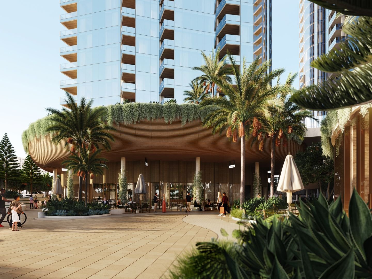Modern architectural plaza with tall glass buildings, palm trees, and green foliage. People are walking and cycling on the spacious tiled area. The structure has a wooden overhang with greenery.