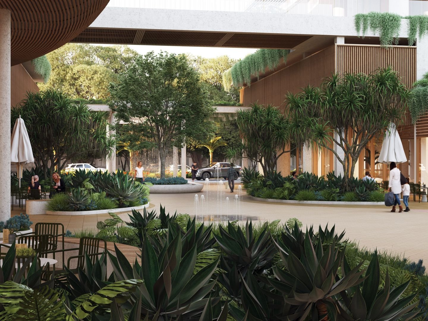 Outdoor courtyard with lush greenery, trees, and seating areas. People walk and sit around a smooth paved area with small fountains. Modern buildings with wooden elements and decorative plants flank the courtyard.