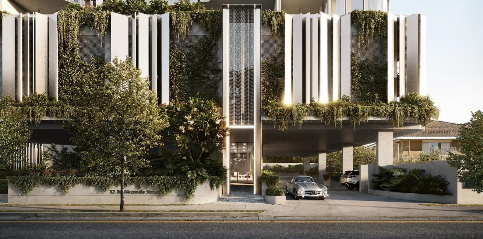 Modern building facade with vertical white panels and lush greenery, two parked cars, and trees in front, located on a city street.