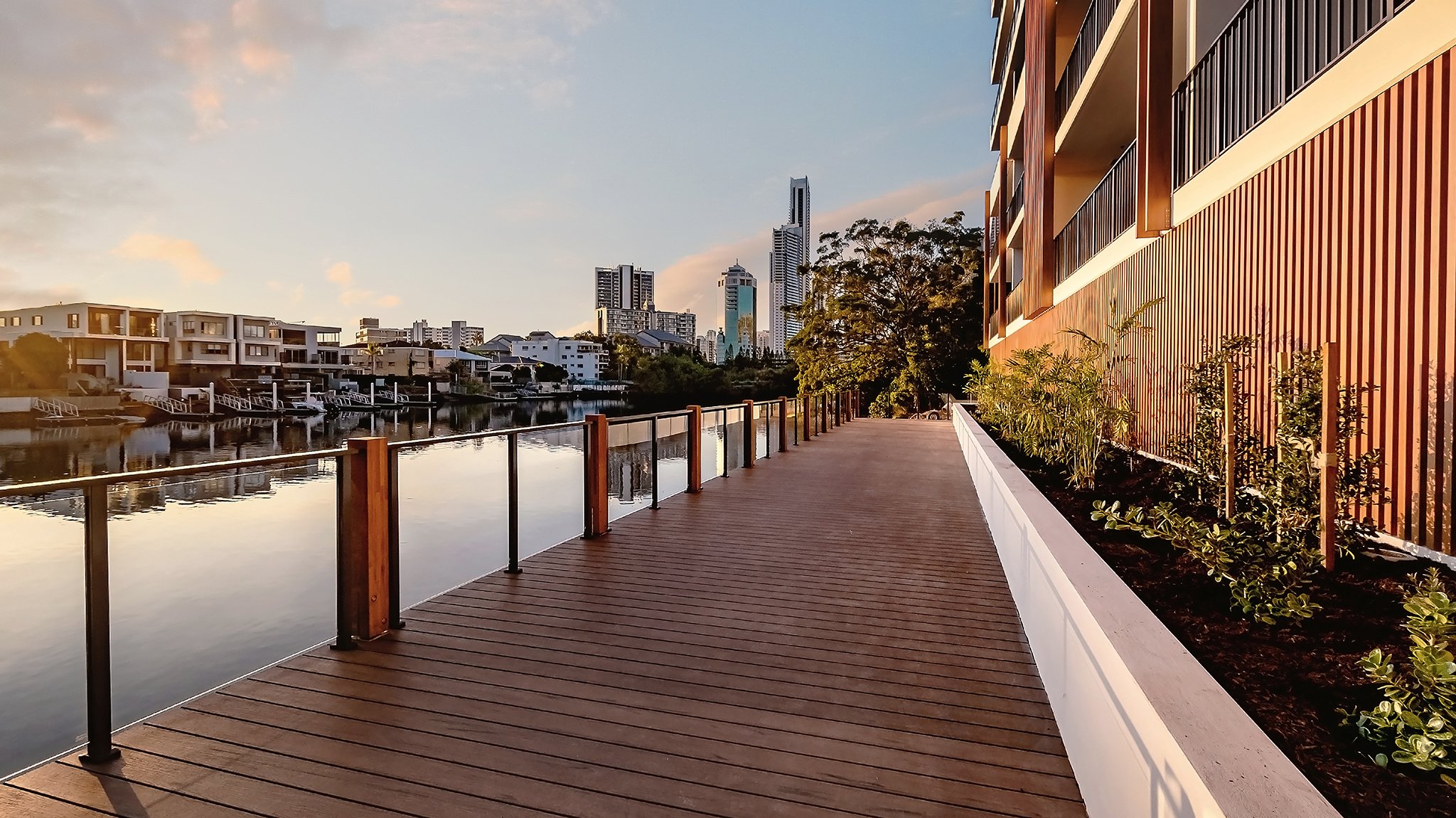 Riverside boardwalk with city skyline and apartment buildings in the background, viewed at sunset.