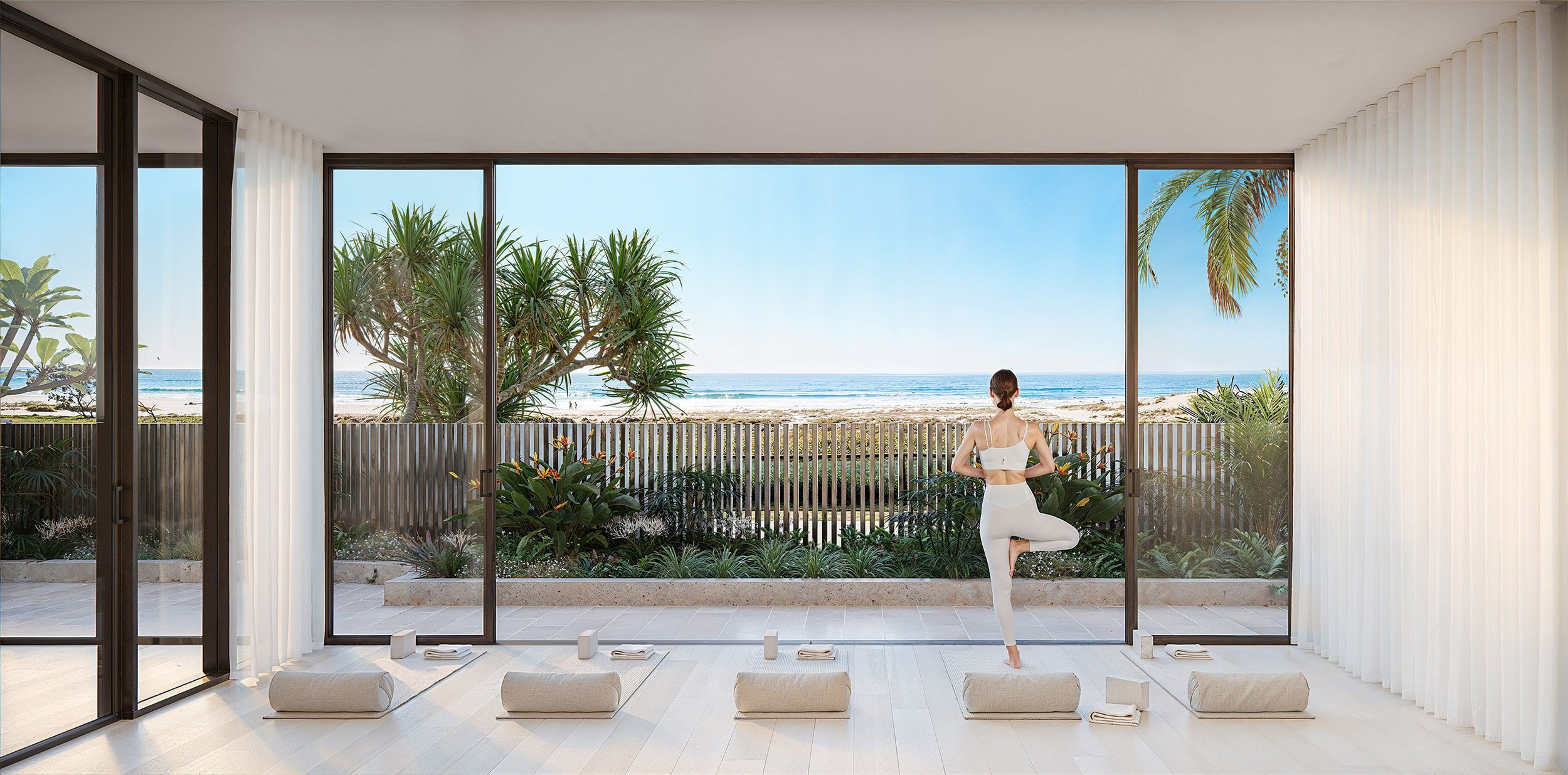 Woman practicing yoga indoors with ocean view through glass walls, surrounded by tropical plants and mats on the floor.