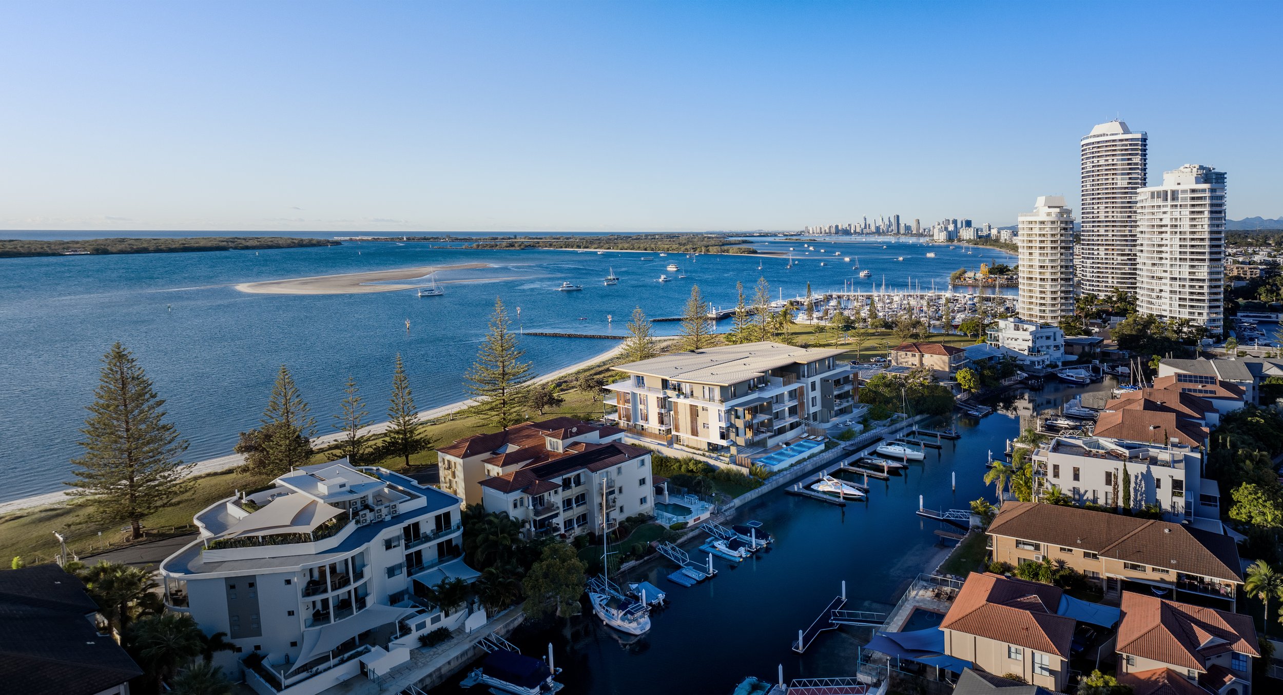 Aerial view of a coastal cityscape featuring high-rise buildings, residential areas, and a marina with boats. The scene includes lush greenery, a sandy beach, and a clear blue sky.
