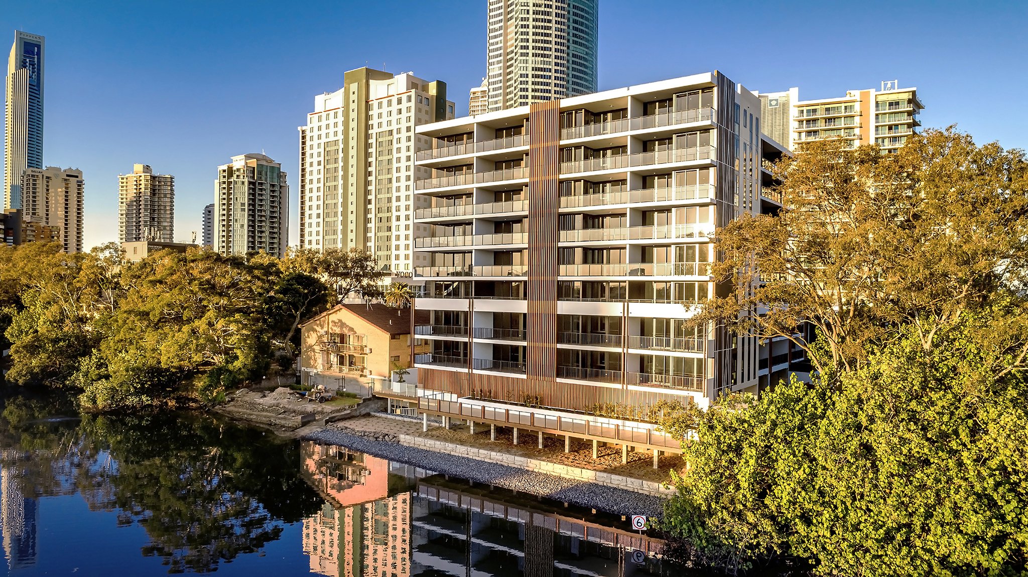 Modern high-rise buildings with a waterfront view, surrounded by trees and reflected in the water, under a clear blue sky.