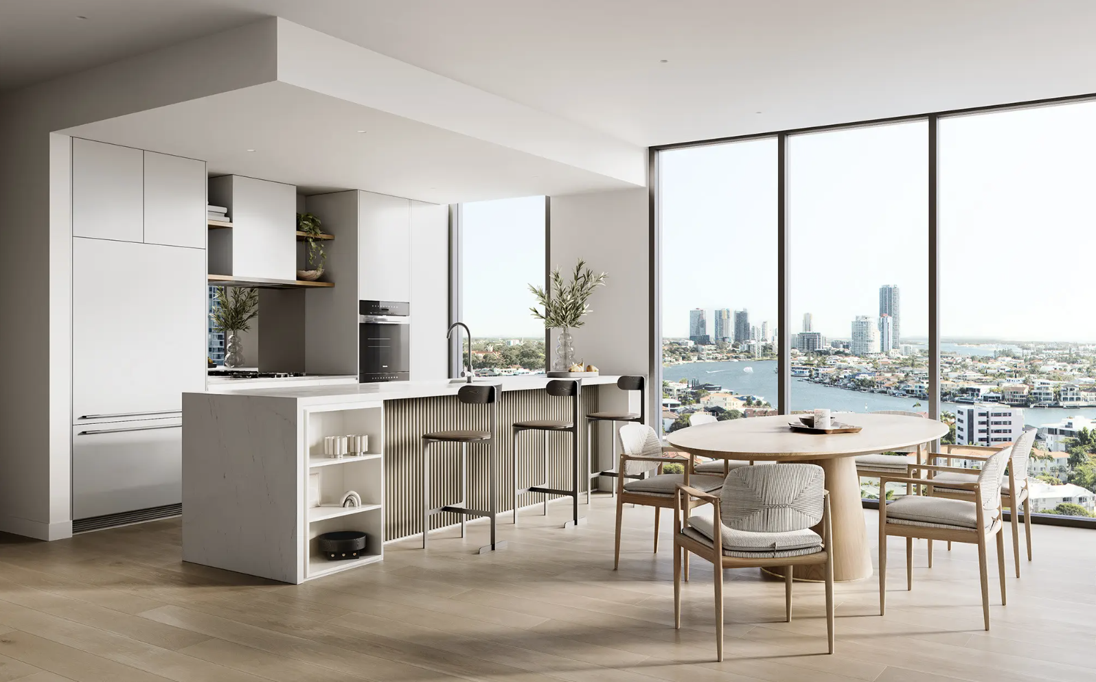 Modern open-plan kitchen and dining area with a view of a cityscape through large windows, featuring minimalist design, a kitchen island with barstools, round dining table with chairs, and light wood flooring.