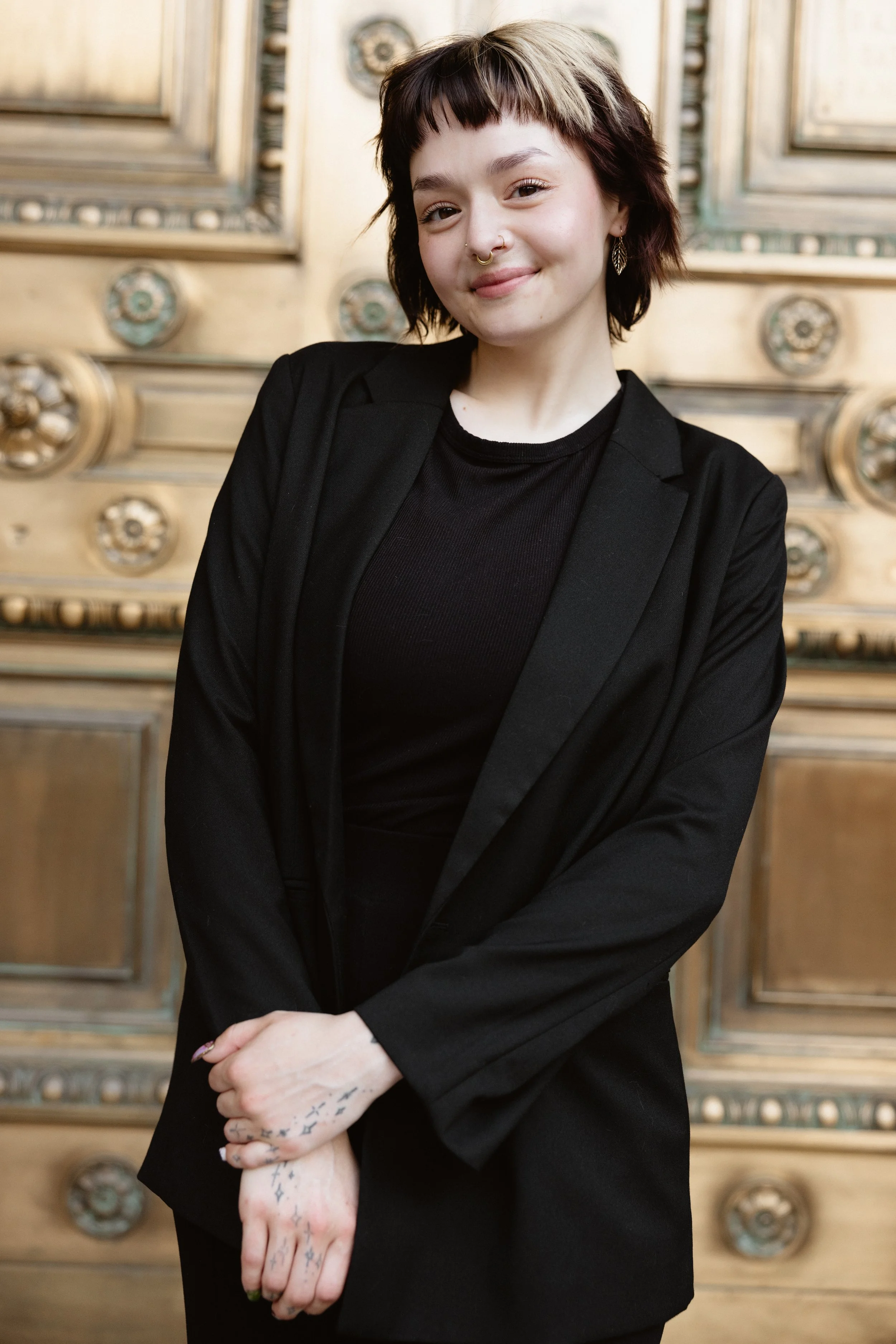 A young woman with short, dark hair with blonde highlights, wearing a black blazer and black shirt, standing in front of a decorative wooden background with ornate carvings and metal accents.
