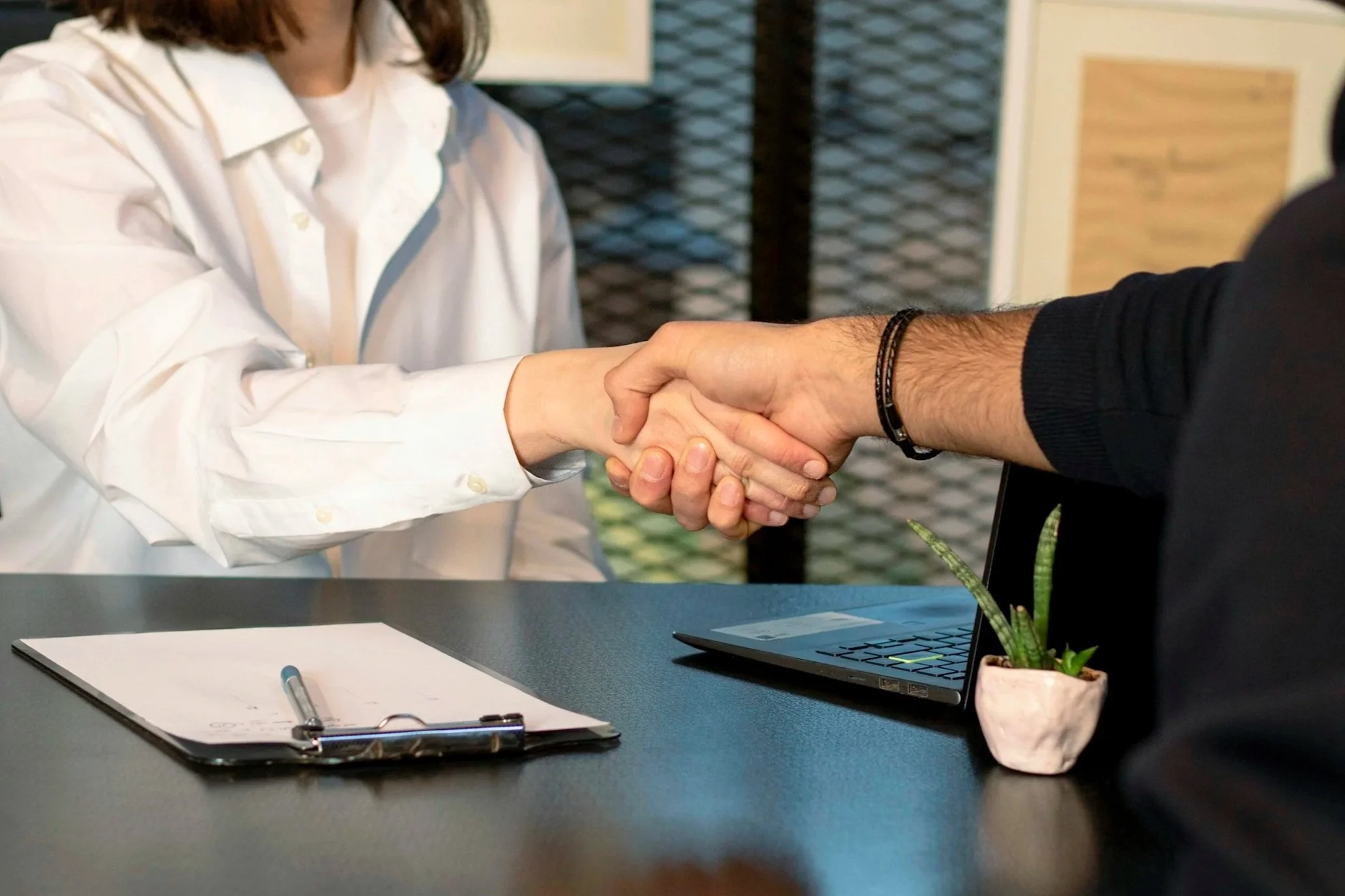 Person wearing a white shirt shaking hands with another person wearing a black shirt in an office setting. On the desk, there is a clipboard with papers, a pen, a laptop, and a small potted plant.