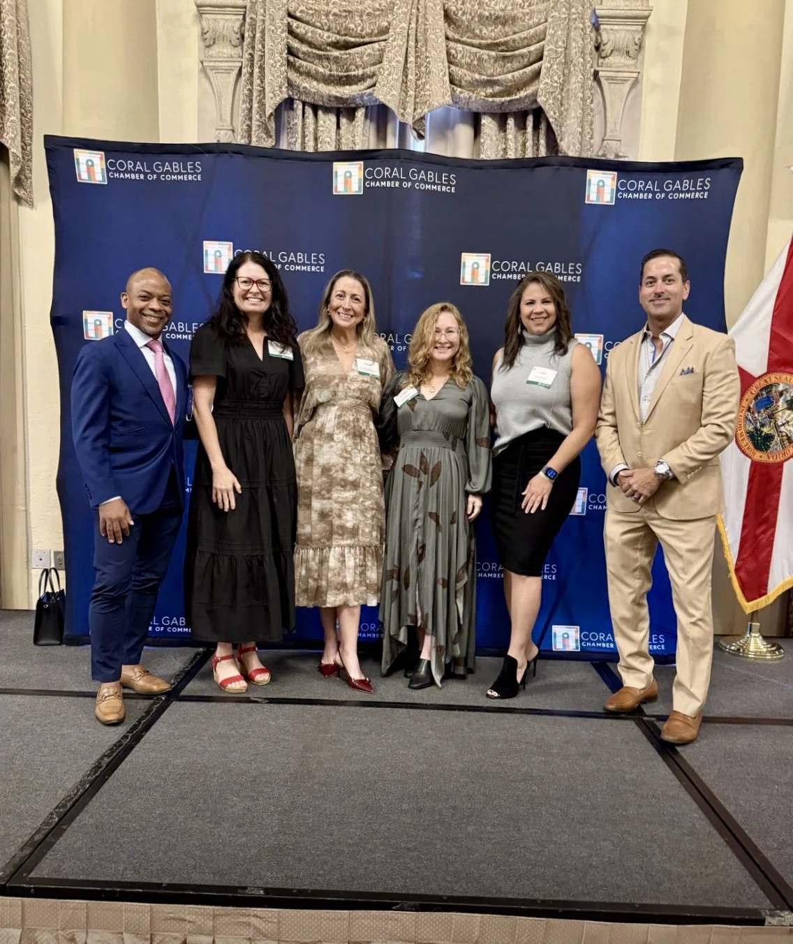 Group of six people standing on a stage in front of a backdrop with the logo of the Coral Gables Chamber of Commerce, posing for a photo. Four women and two men, dressed in business attire, smiling.