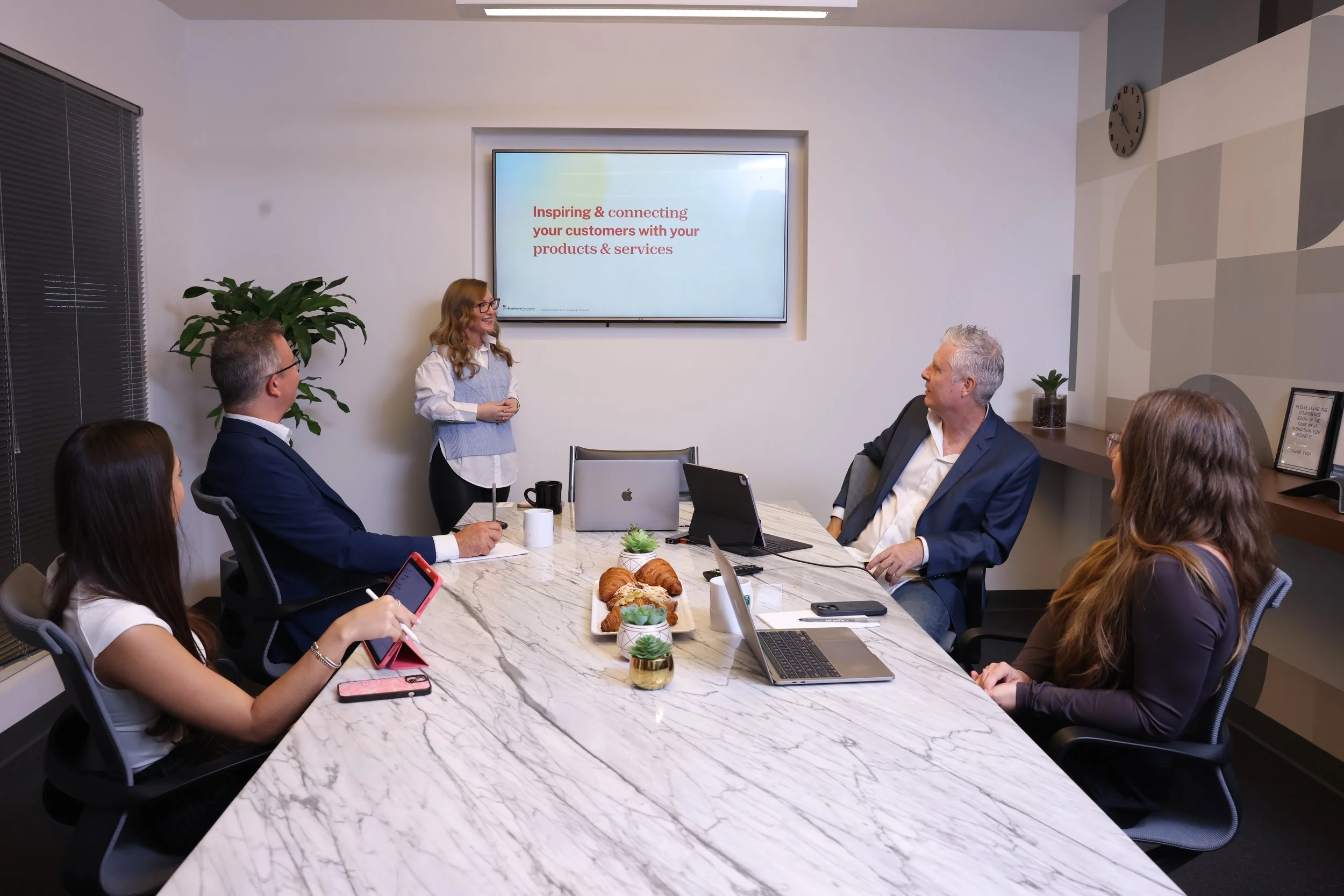 A woman presents in a conference room with six people seated around a marble table, watching her. The presentation screen displays the text 'Inspiring & connecting your customers with your products & services.' There are laptops, a smartphone, a coffee mug, pastries, and small potted plants on the table. The room has white walls, a large window with blinds, and decorative plants.