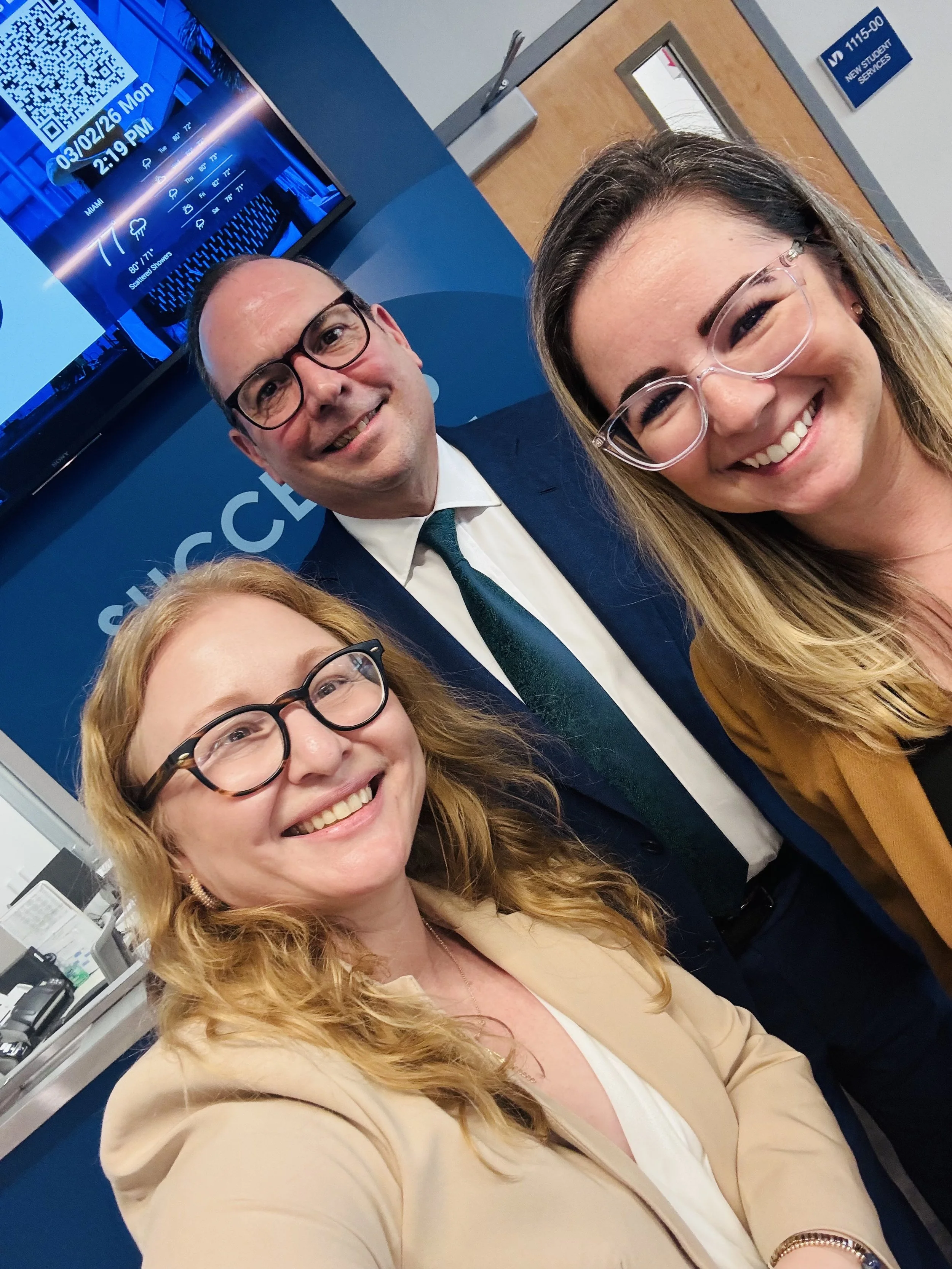 A group of four people smiling for a selfie in an office or reception area. There are two women in the foreground and two men in the background, with a computer monitor displaying the date and time on the wall behind them.