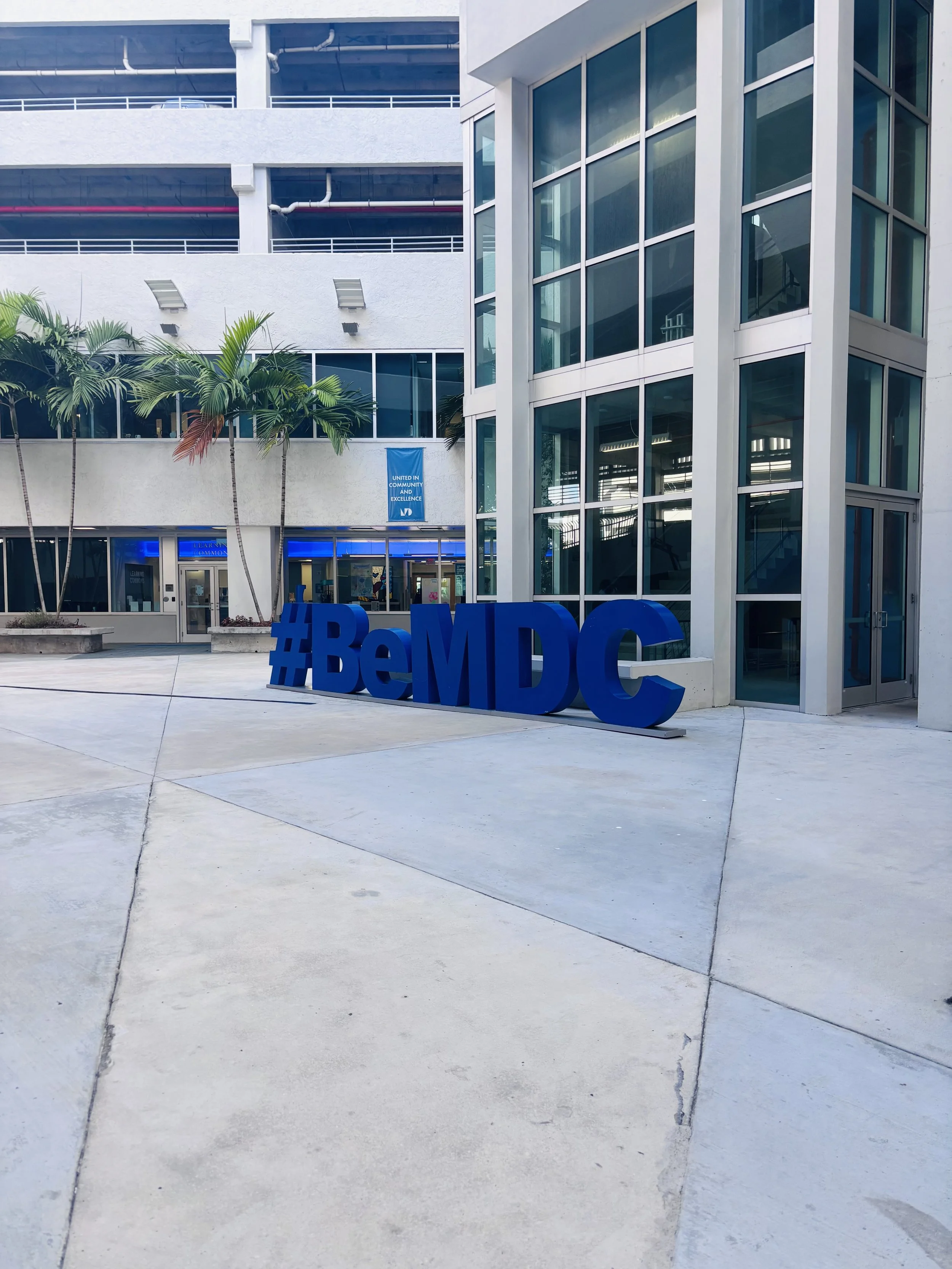 Exterior view of a modern building with large glass windows, palm trees, and a blue hashtag sign reading #BeMDC in the foreground.