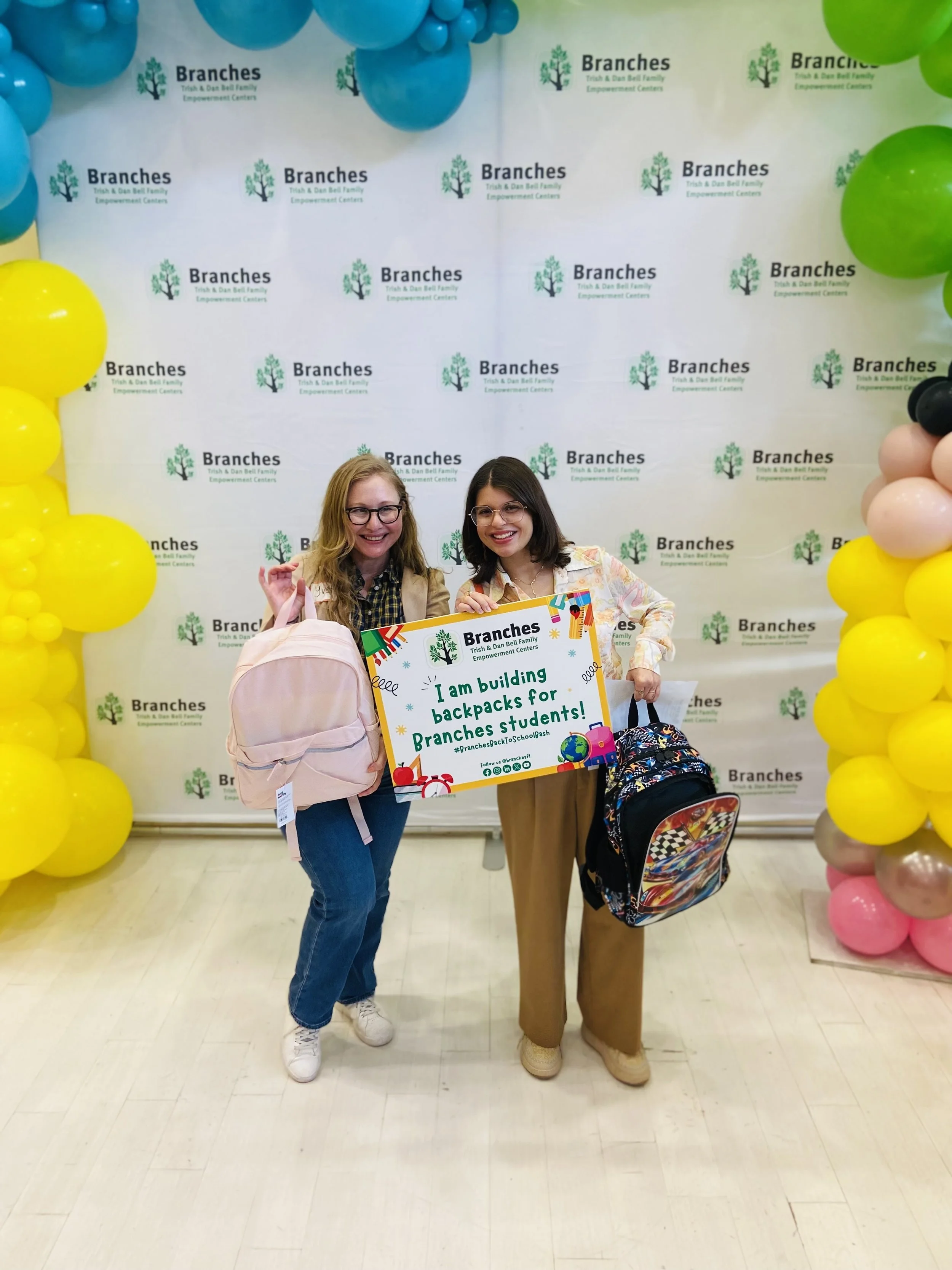 Two young women smiling and posing with backpacks at a school supply donation event. They are holding a colorful sign that says they are building backpacks for students, standing in front of a backdrop with balloons on the sides and a logo that reads