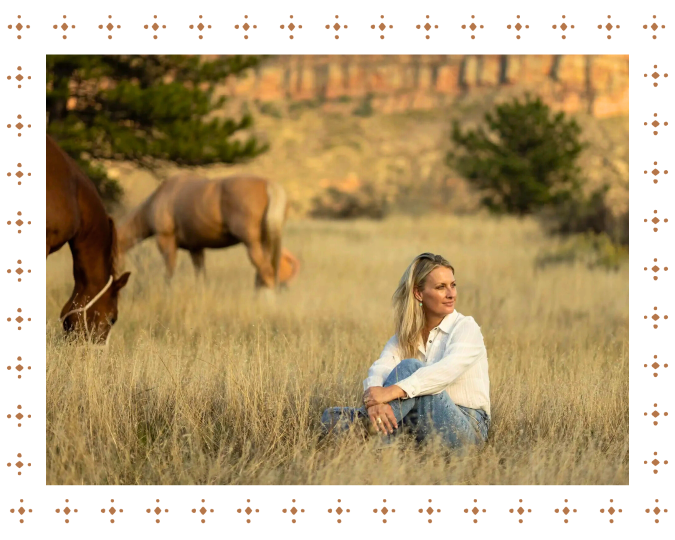 A woman sitting on the grass in a field with two grazing horses, trees, and a red rock canyon in the background during sunset.