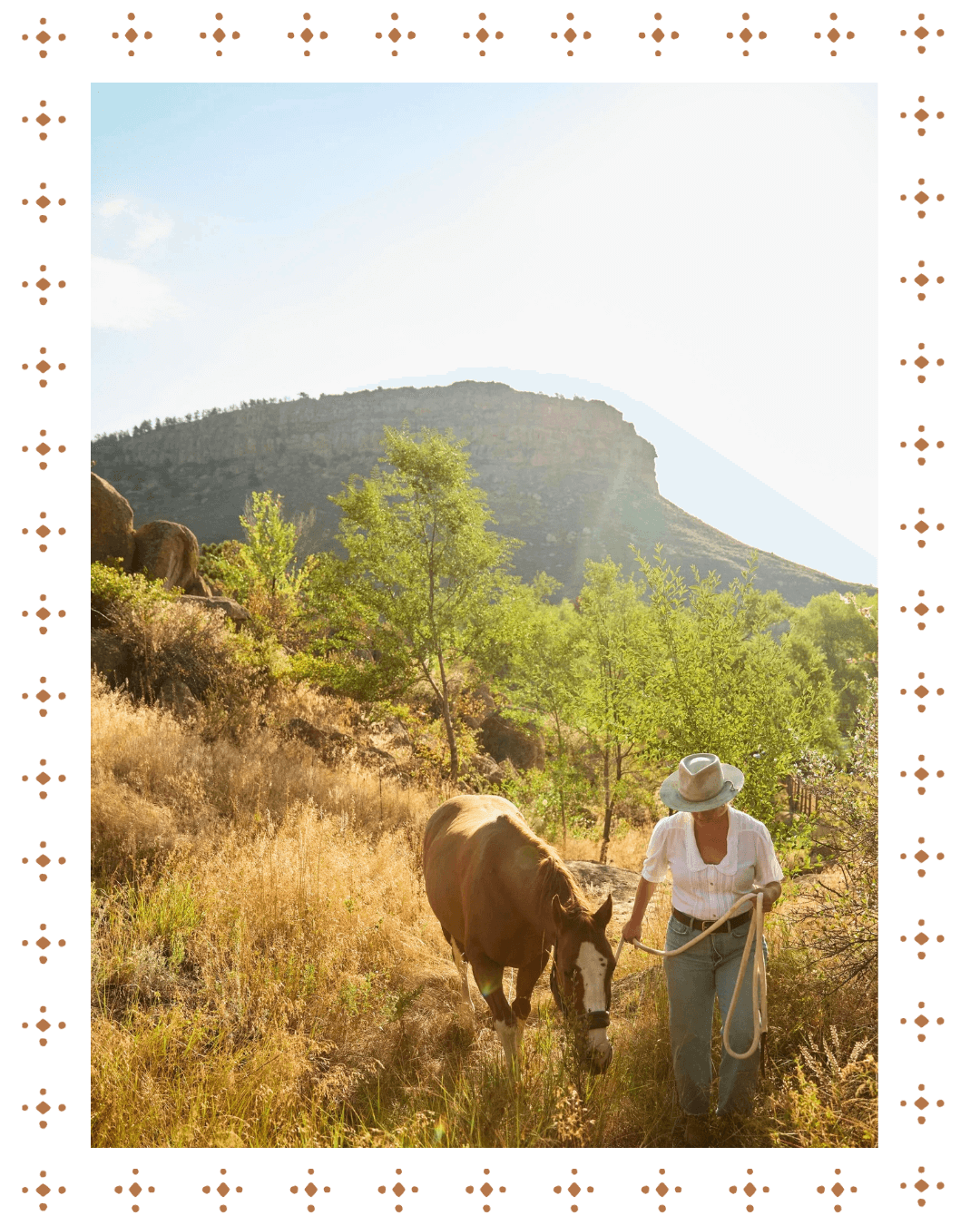 A woman in a white shirt and wide-brimmed hat walks a brown and white horse through a grassy field with green trees, mountains, and a bright sky in the background.