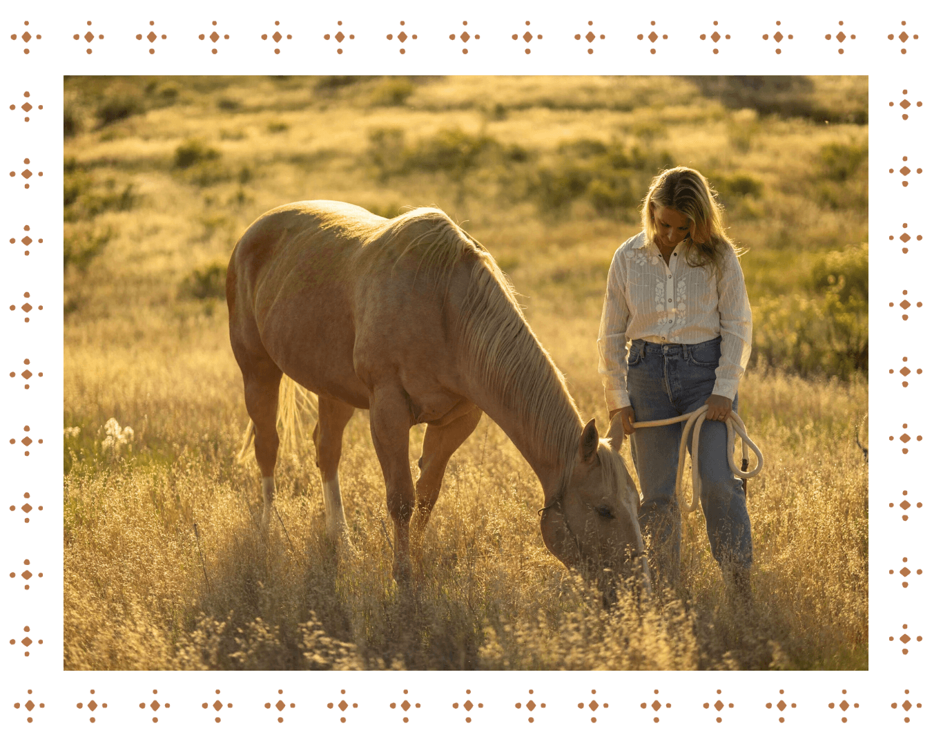 A woman in a white blouse and jeans holding a lead rope walks with a light brown horse in a grassy field during sunset.