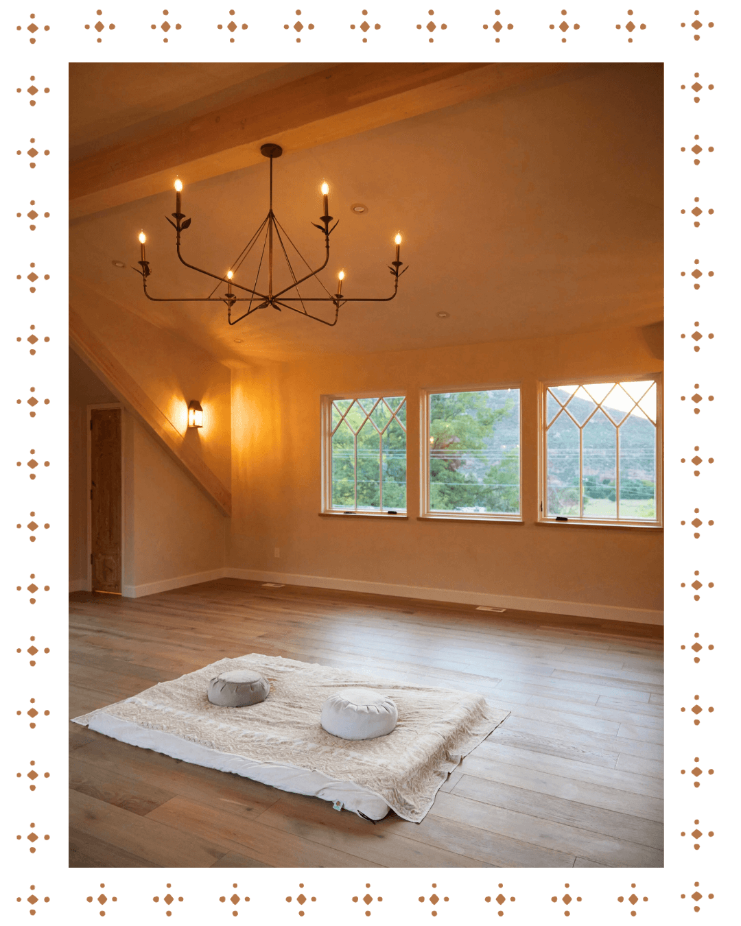 Interior of a spacious room with wood flooring, a large window with a scenic view, a chandelier hanging from the ceiling, and two floor cushions on a rug on the floor.
