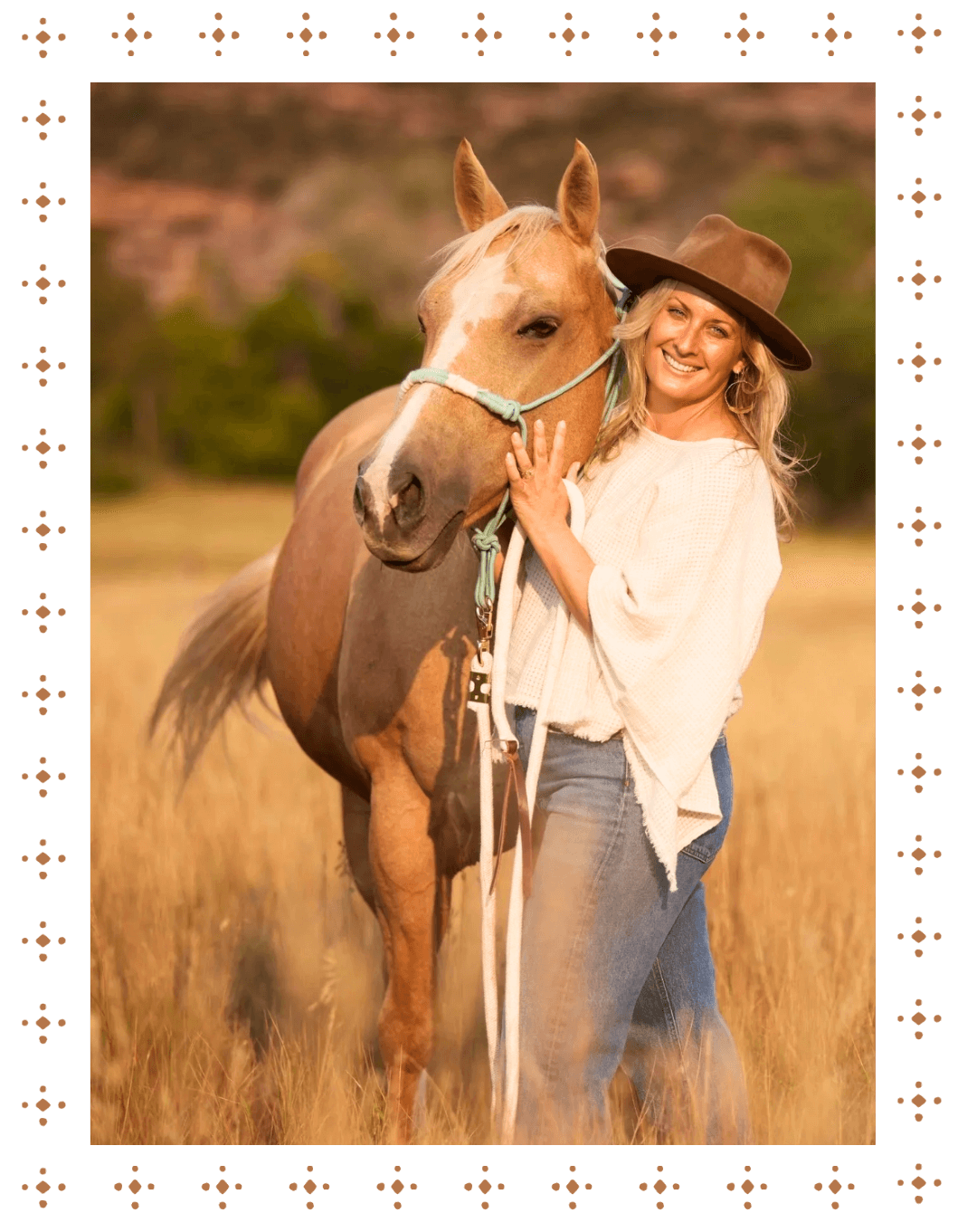 A woman in a white sweater and jeans smiling and standing next to a light brown horse in an outdoor field, with trees in the background.