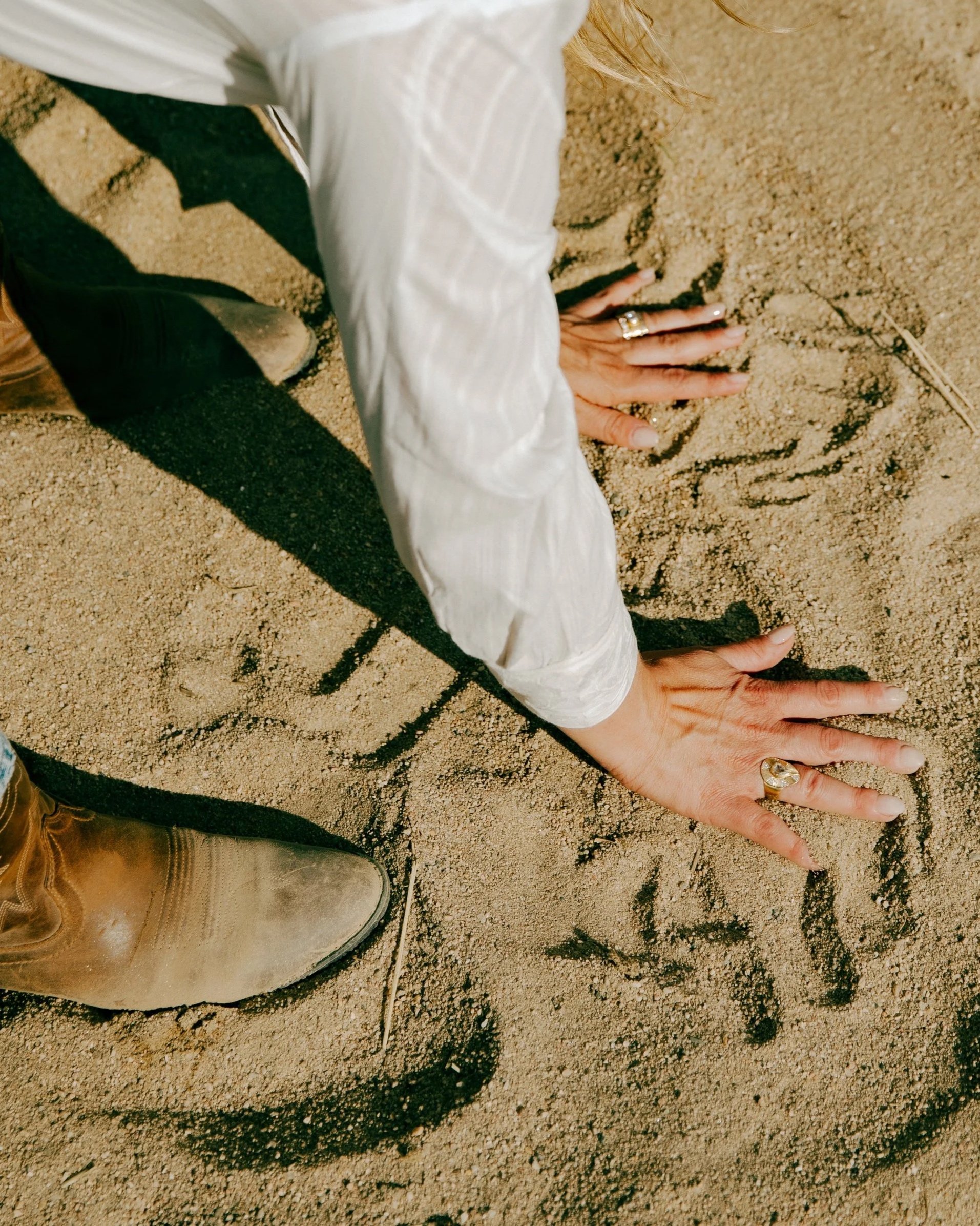 Person's hands and part of shoe on sandy ground, writing or drawing in the sand with a finger.