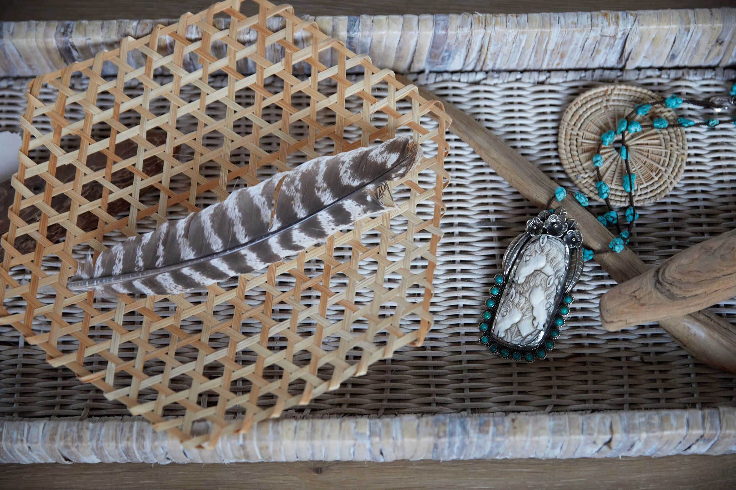 A woven basket containing a striped feather, a silver and turquoise jewelry pendant, a turquoise beaded necklace, and a piece of driftwood, all on a wicker surface.