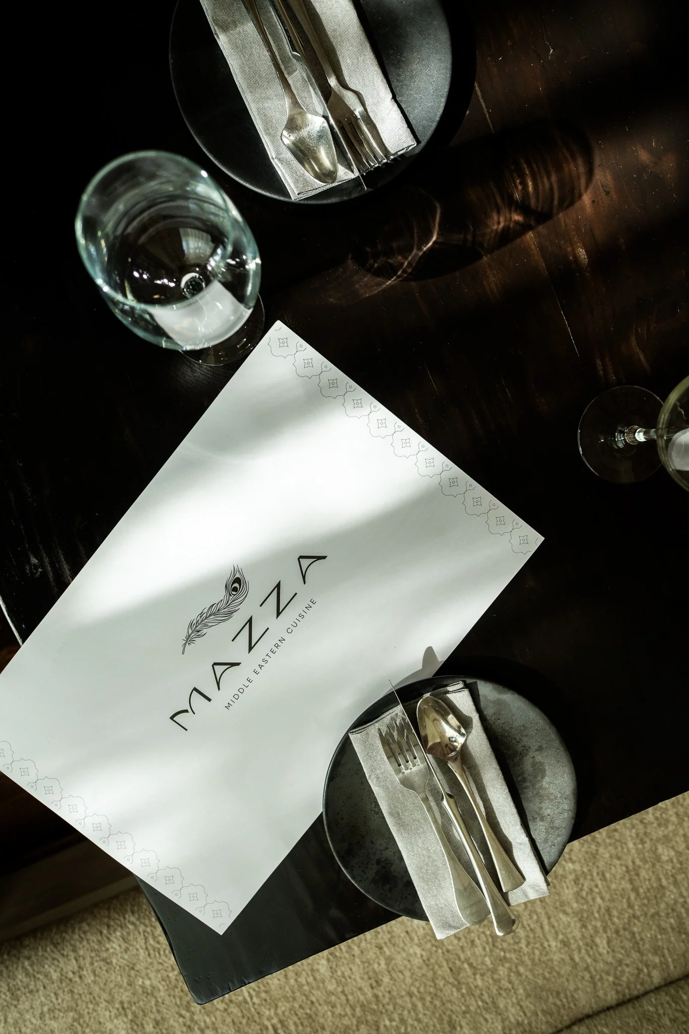 A restaurant table setting with a black plate, a fork, a knife, and a spoon on a folded napkin, a glass of water, and a menu on a dark wooden table.