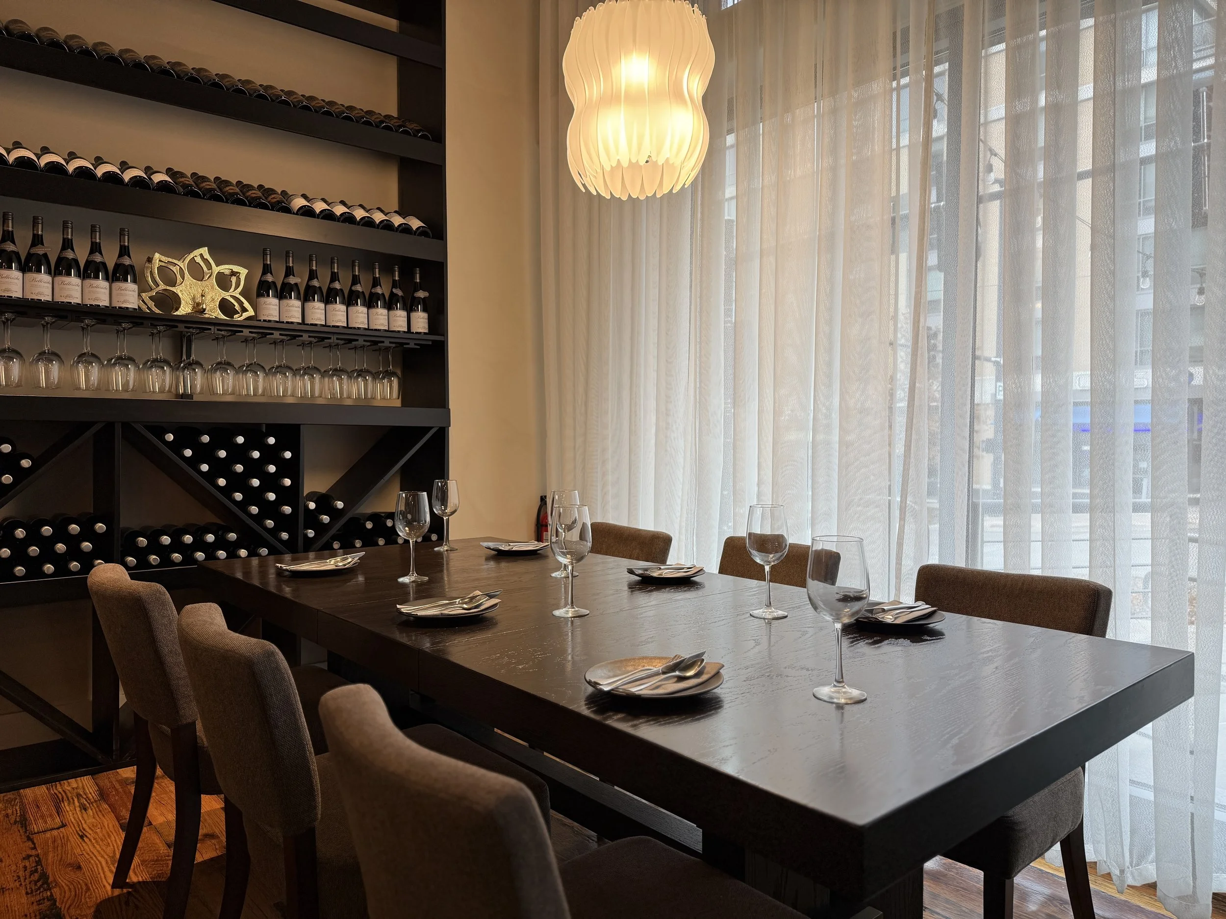 A modern dining room with a long dark wooden table set with glassware and napkins. There are several chairs around the table, and a large window with sheer curtains letting in natural light. A wall-mounted wine rack with wine bottles and glasses is visible, along with a decorative piece on the shelf. A hanging light fixture illuminates the room.