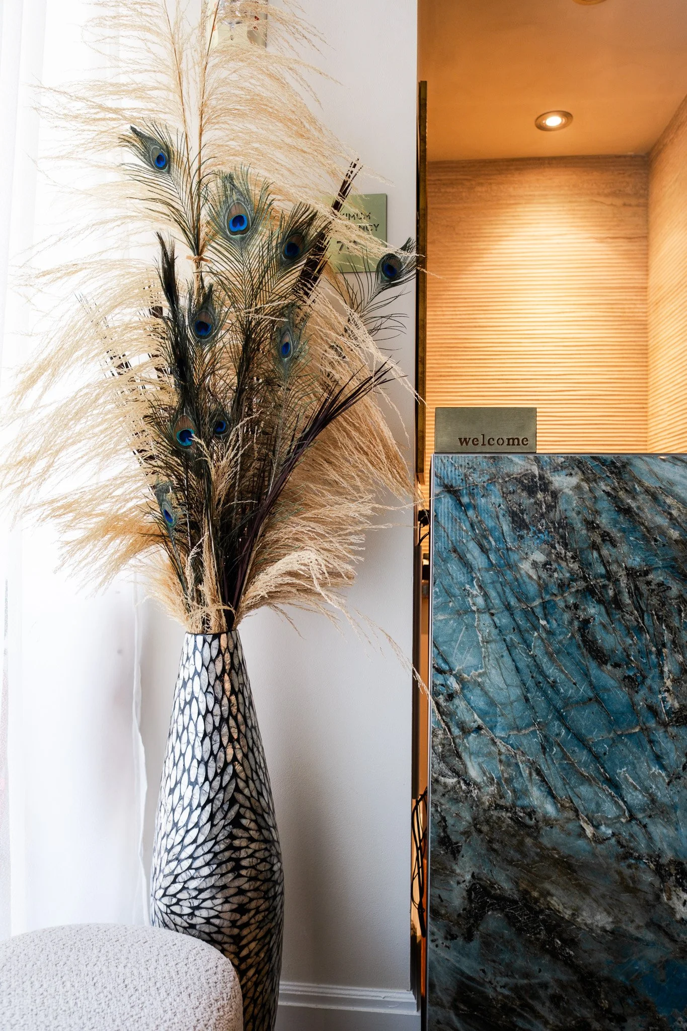 A tall vase with a mosaic pattern holds a bouquet of peacock feathers and dried pampas grass. In the background, there is a sign with the word "welcome" on it, and a textured, wooden wall with recessed lighting. To the left, part of a white textured stool is visible.