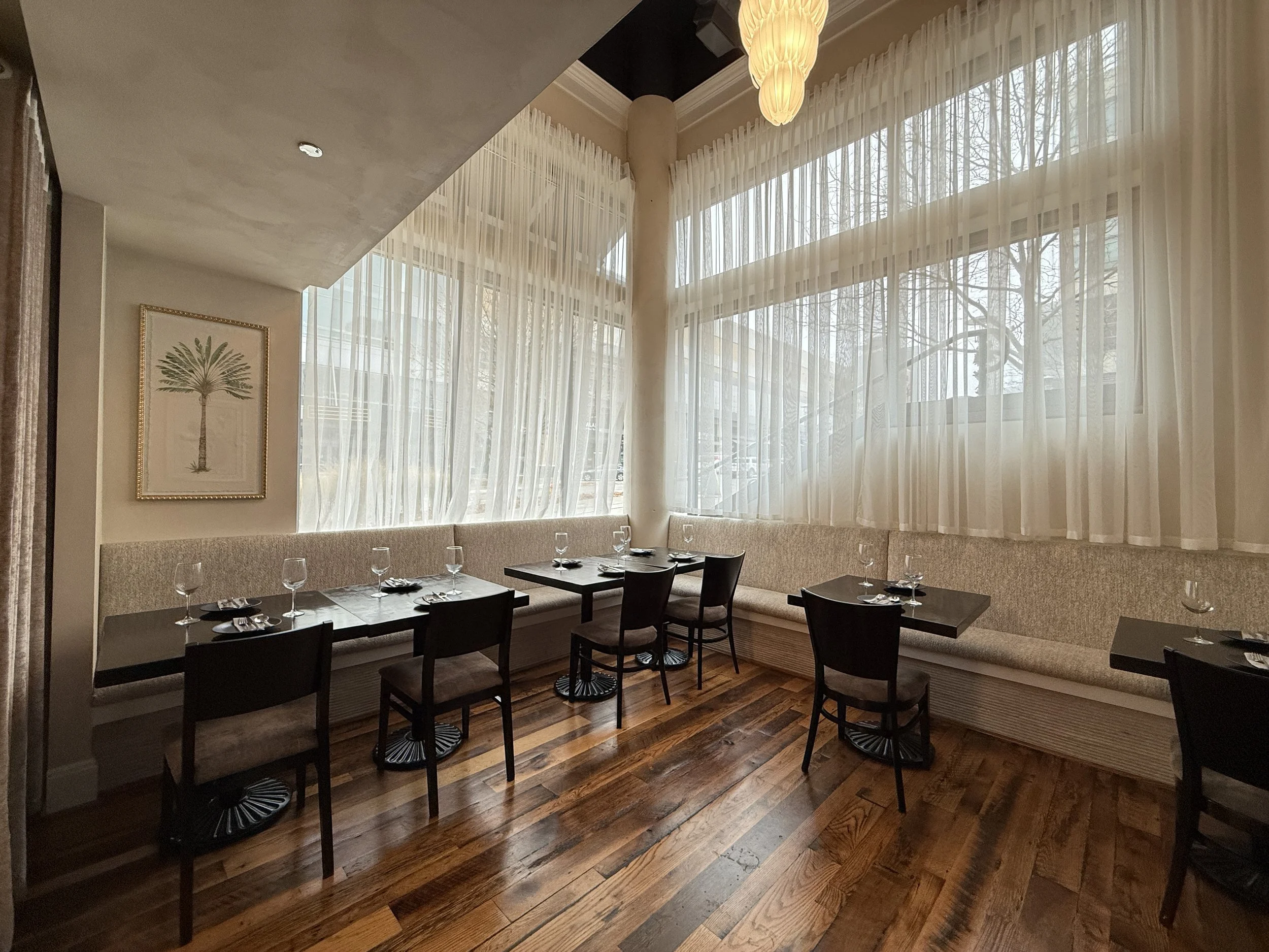 Interior of a restaurant with large windows, sheer curtains, black tables set with glasses and silverware, and black chairs. There is a framed botanical print on the wall and a hanging light fixture.