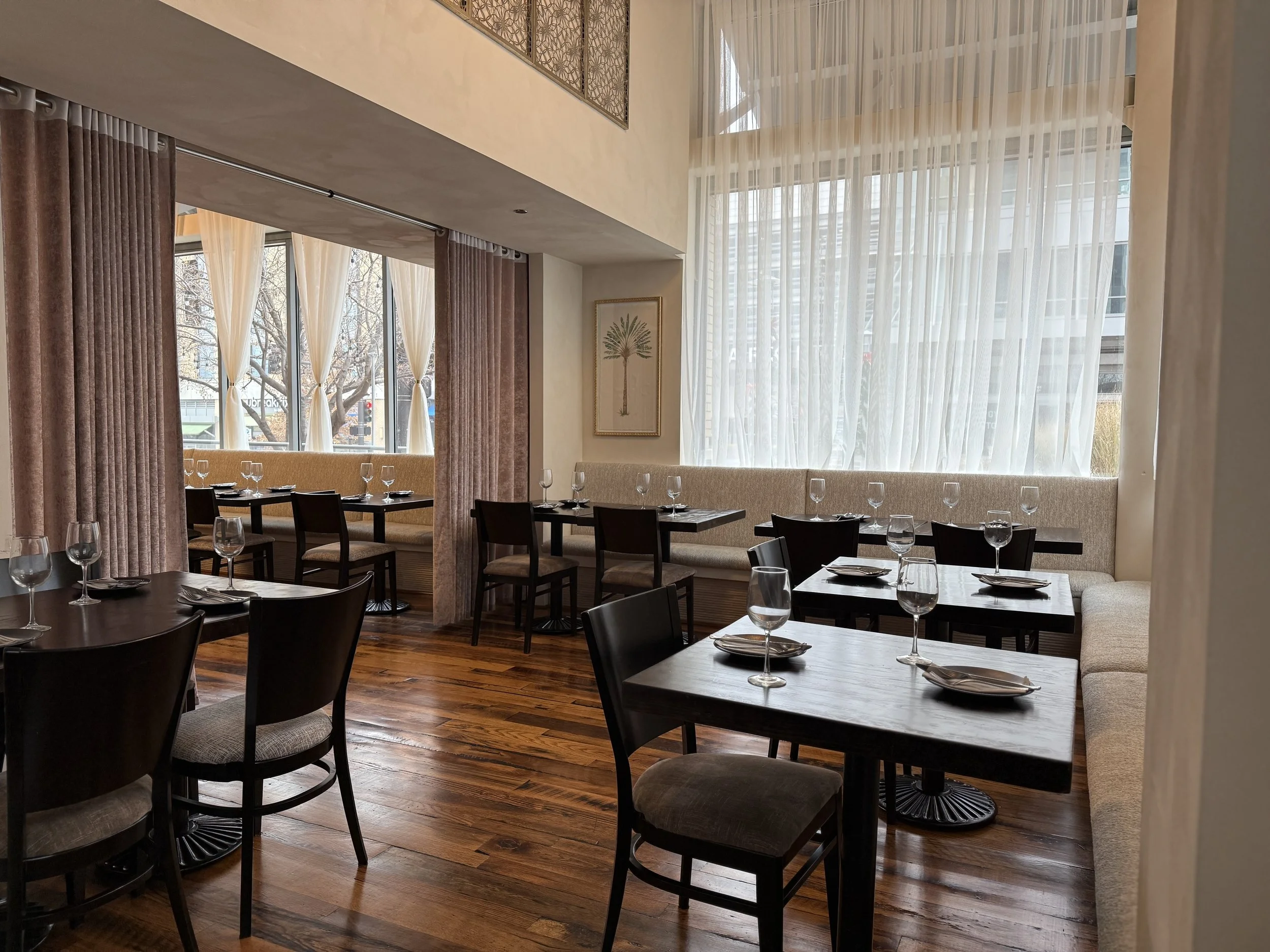 Interior of a restaurant with black tables set with plates and glasses, wooden floors, large windows with sheer white curtains, and a framed botanical picture on the wall.