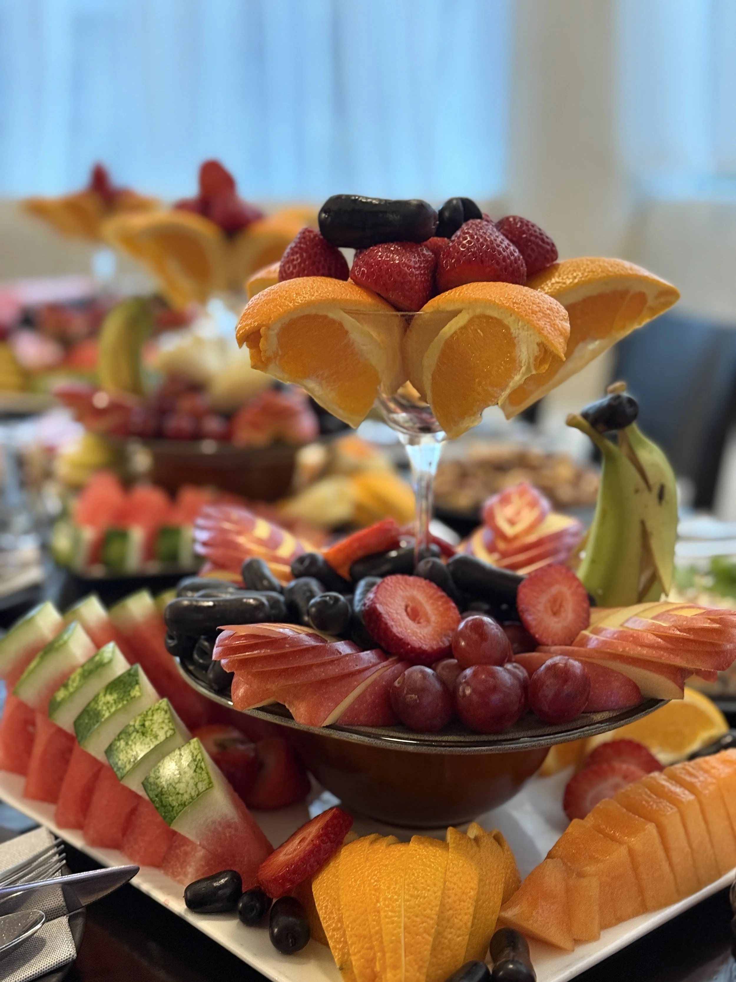 A tiered platter of assorted fresh sliced fruits including strawberries, oranges, watermelon, honeydew, cantaloupe, mango, and grapes.