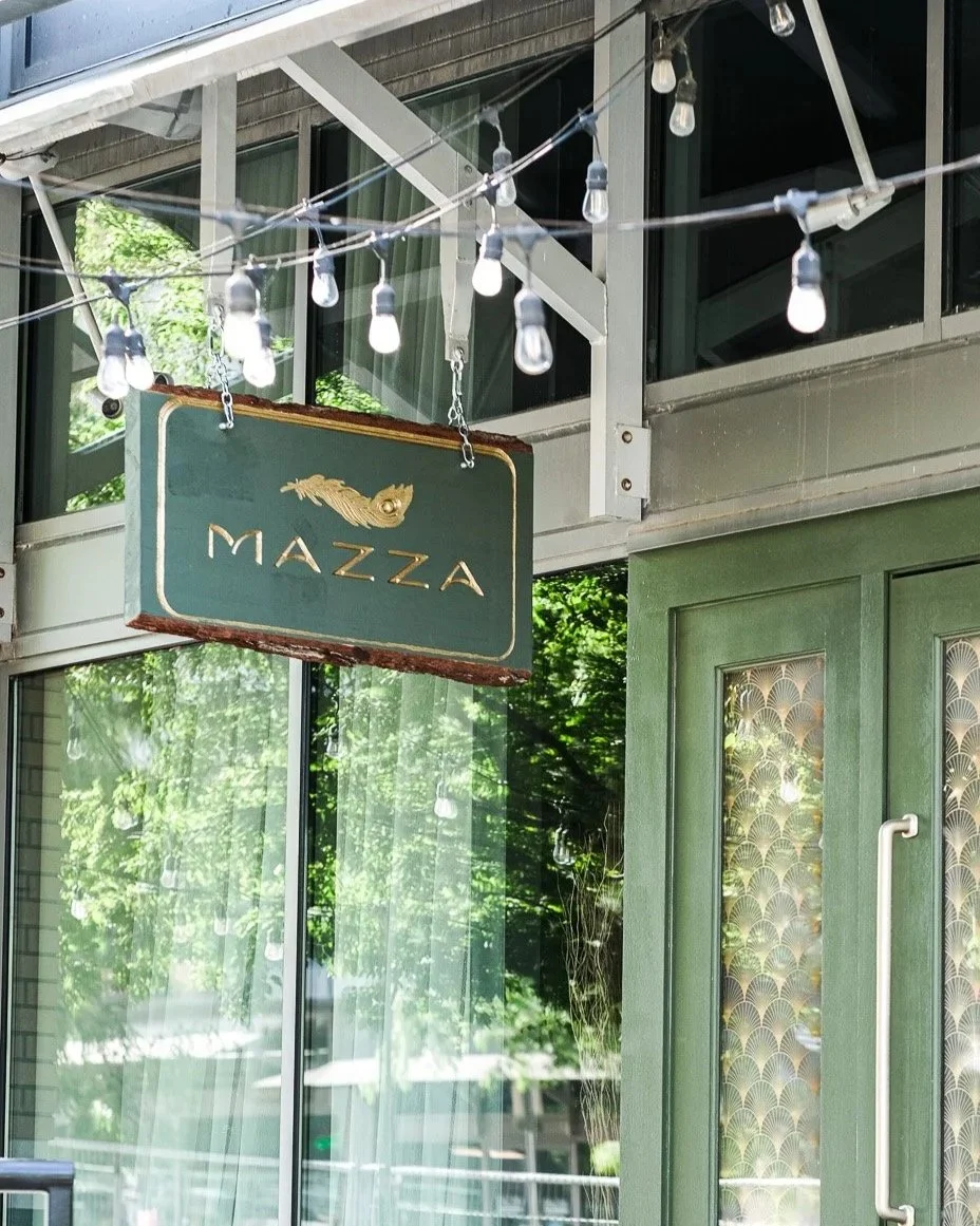 A green sign with gold text that says 'MAZZA' hanging outside a building under string lights, with glass windows reflecting green trees.