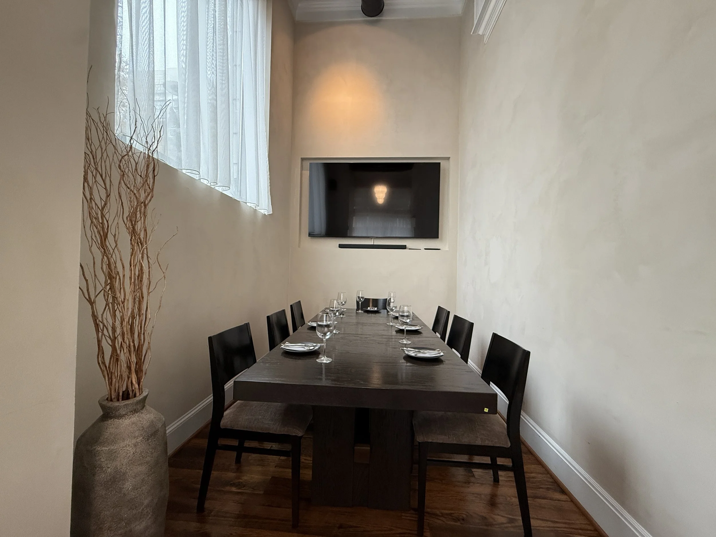 Dining room with a long wooden table set with wine glasses and silverware, surrounded by black chairs, and a large television mounted on the wall. A tall vase with dried branches is placed on the floor near a window with sheer curtains, and the room has light-colored walls and dark hardwood floors.