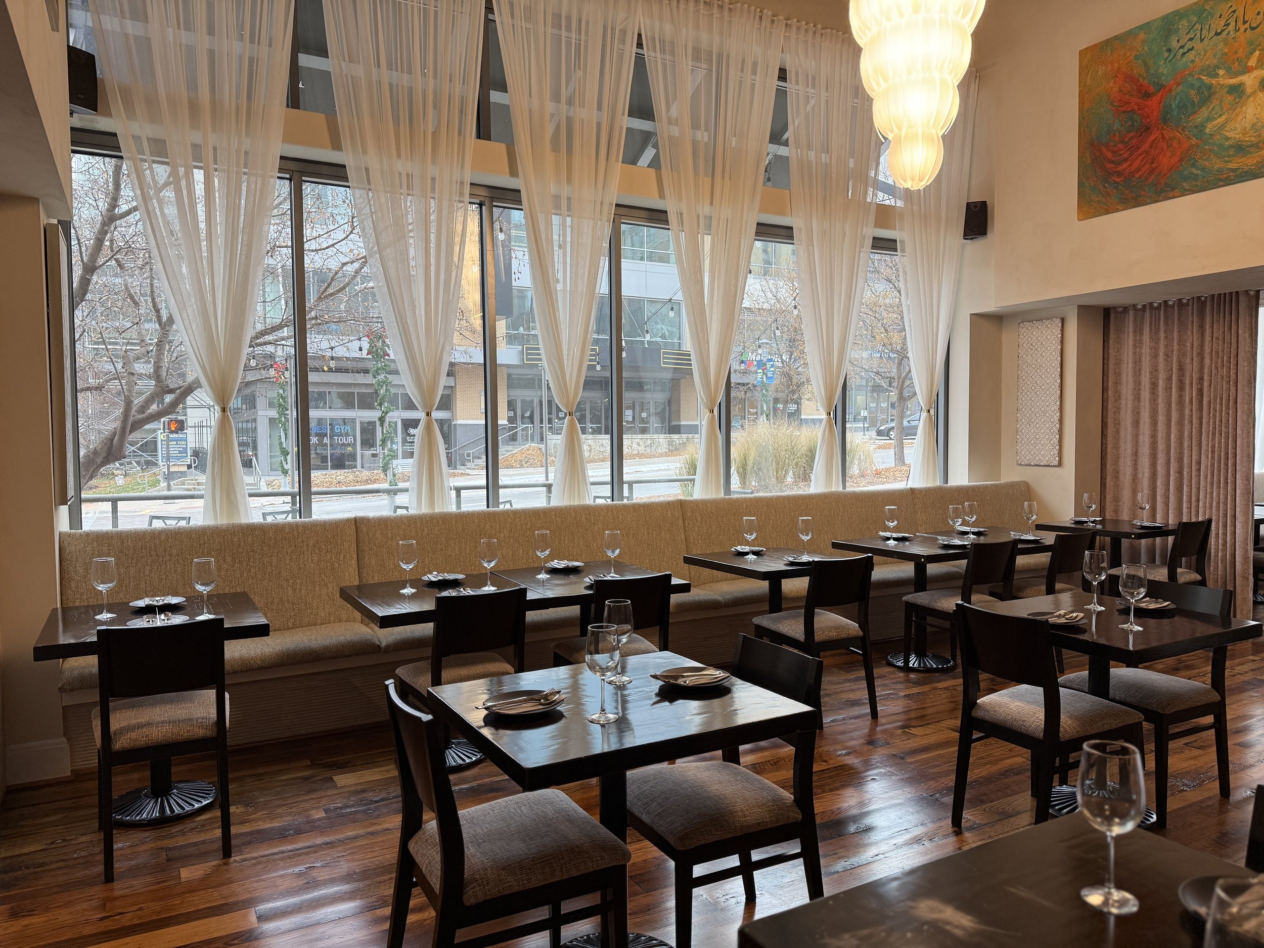 Interior of a restaurant with large windows, sheer curtains, and tables set with wine glasses, forks, and knives.