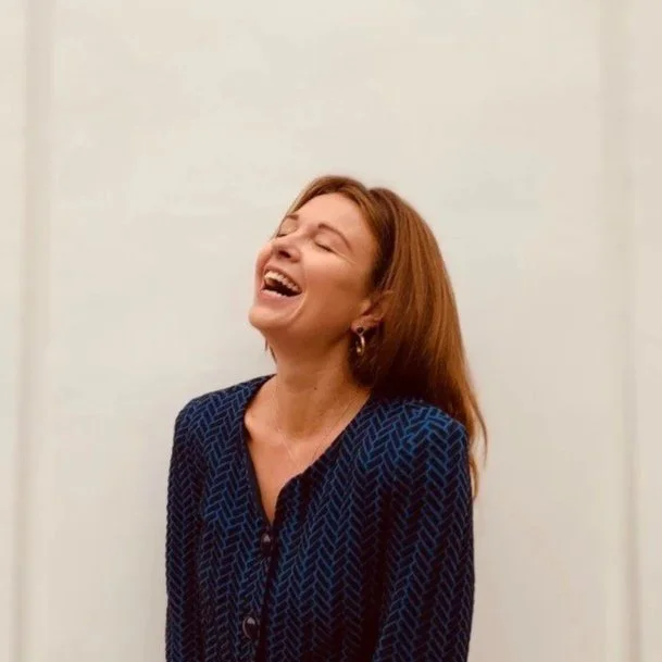 A woman with red hair laughing with her eyes closed, wearing a navy blue patterned top, standing against a plain white wall.