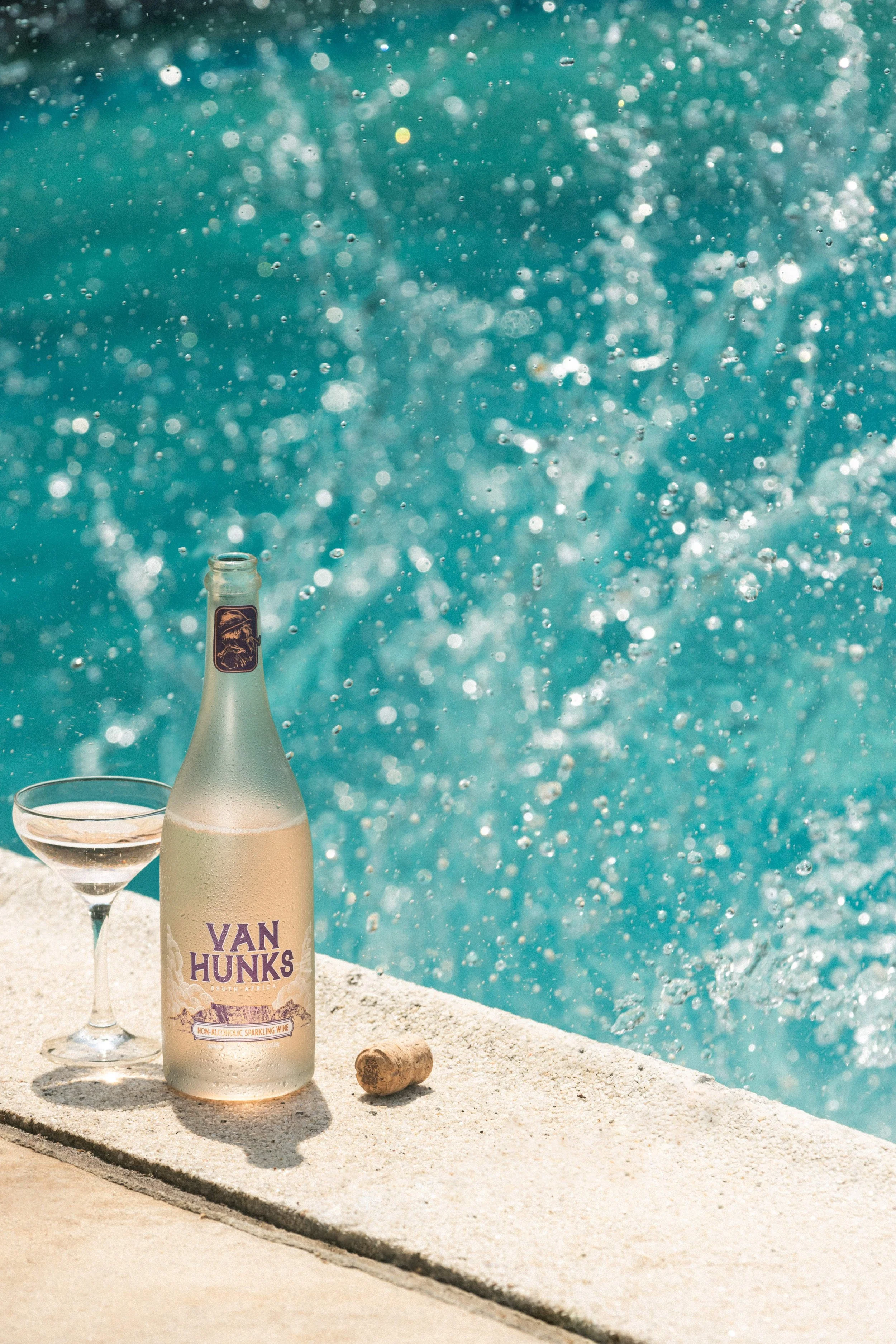 A bottle of Van Hunks sparkling non-alcoholic wine, a wine glass, and a cork on a poolside edge with water splashing in the background.
