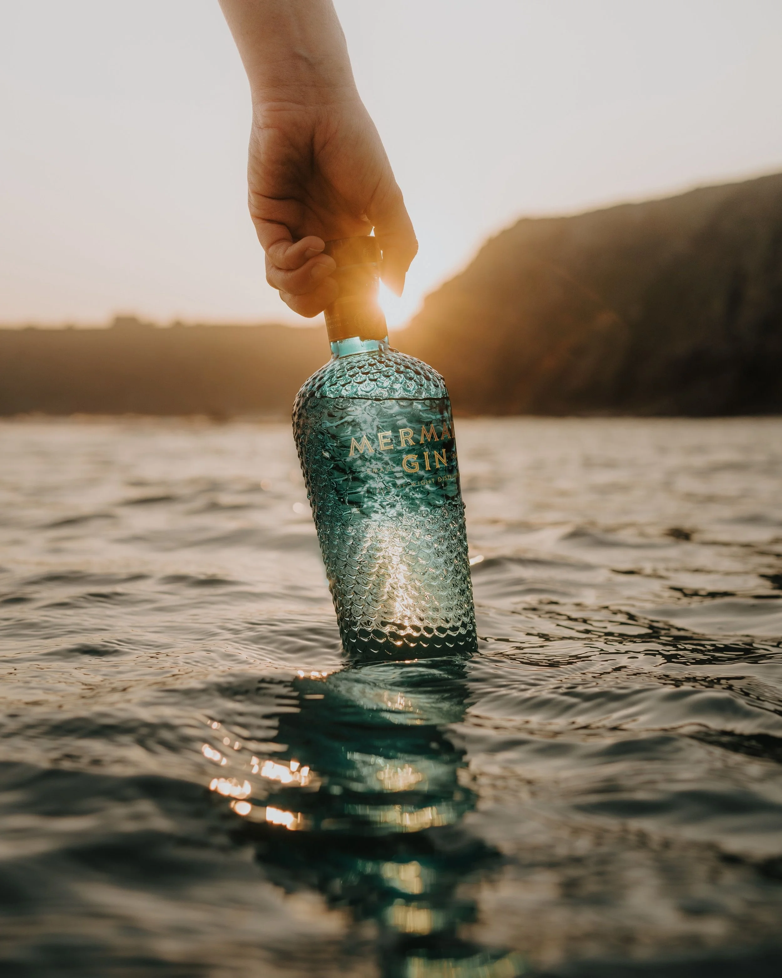 A hand holding a bottle of Mermaids Gin partially submerged in water during sunset, with a distant landscape in the background.