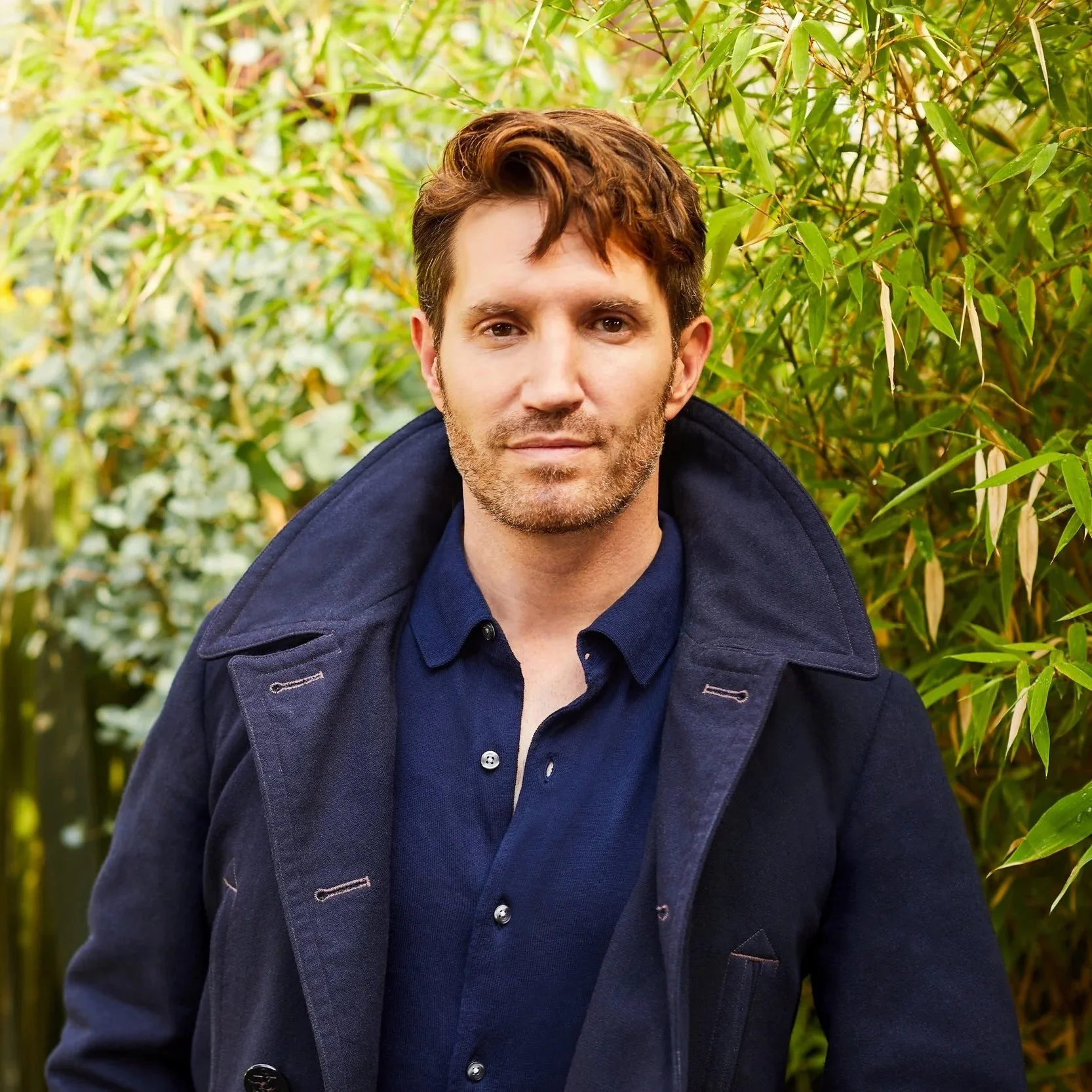 A man with brown hair and facial hair wearing a navy blue shirt and coat stands outdoors in front of green foliage.