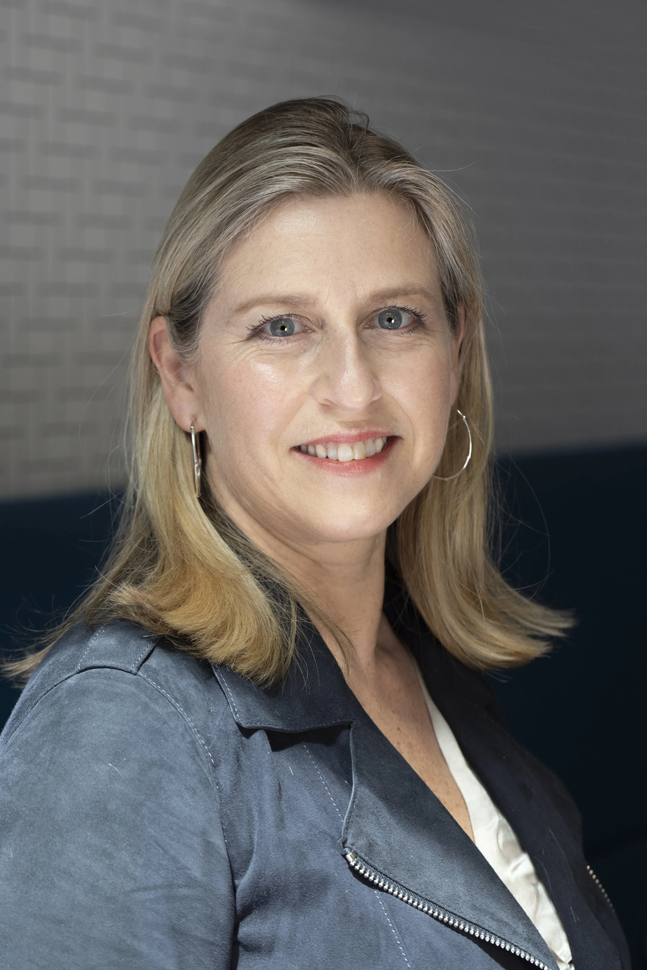 Portrait of a woman with blonde hair wearing hoop earrings and a dark jacket, smiling at the camera.