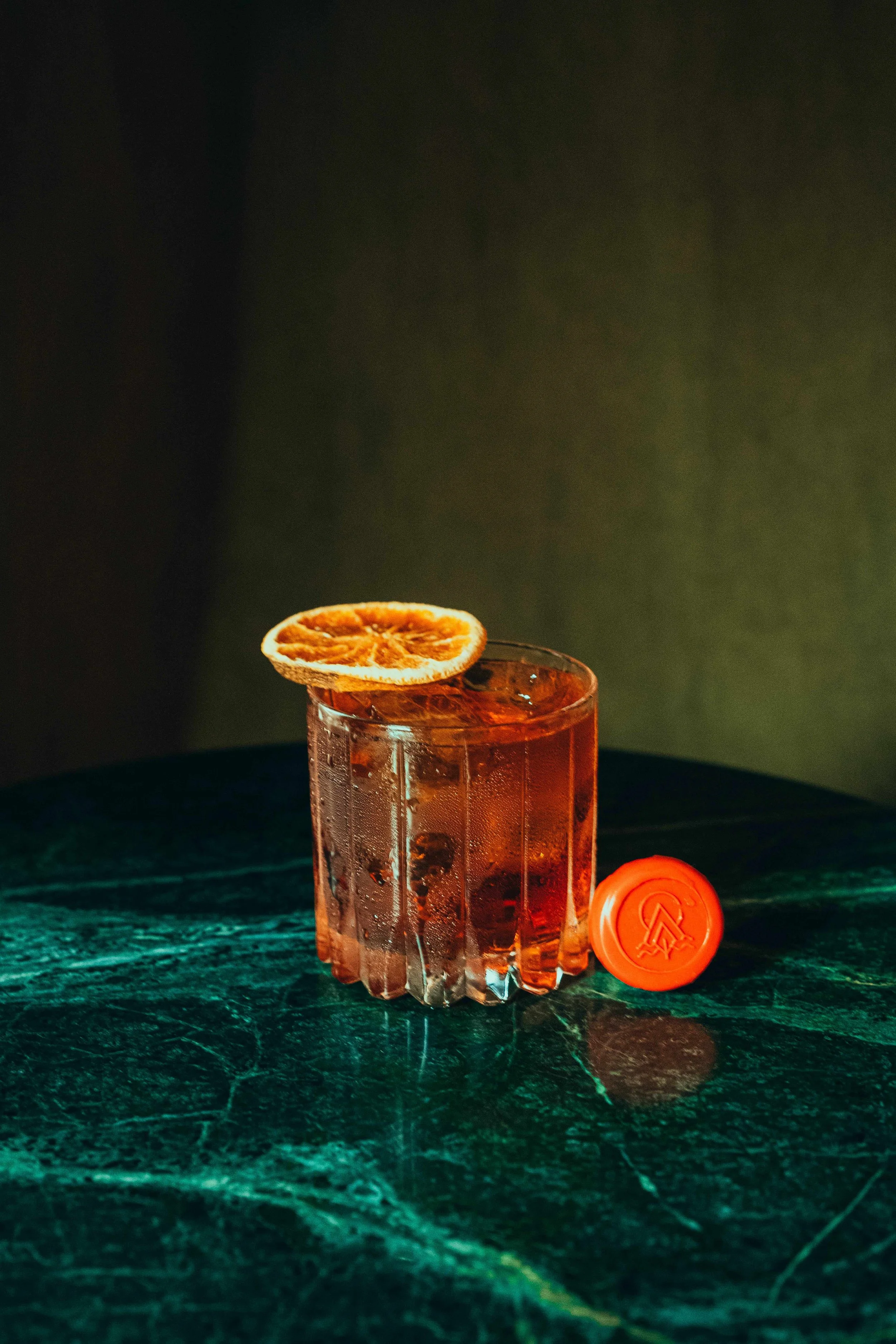 A glass of brown cocktail garnished with a dried orange slice, placed on a dark green marble surface with an orange bottle cap nearby.