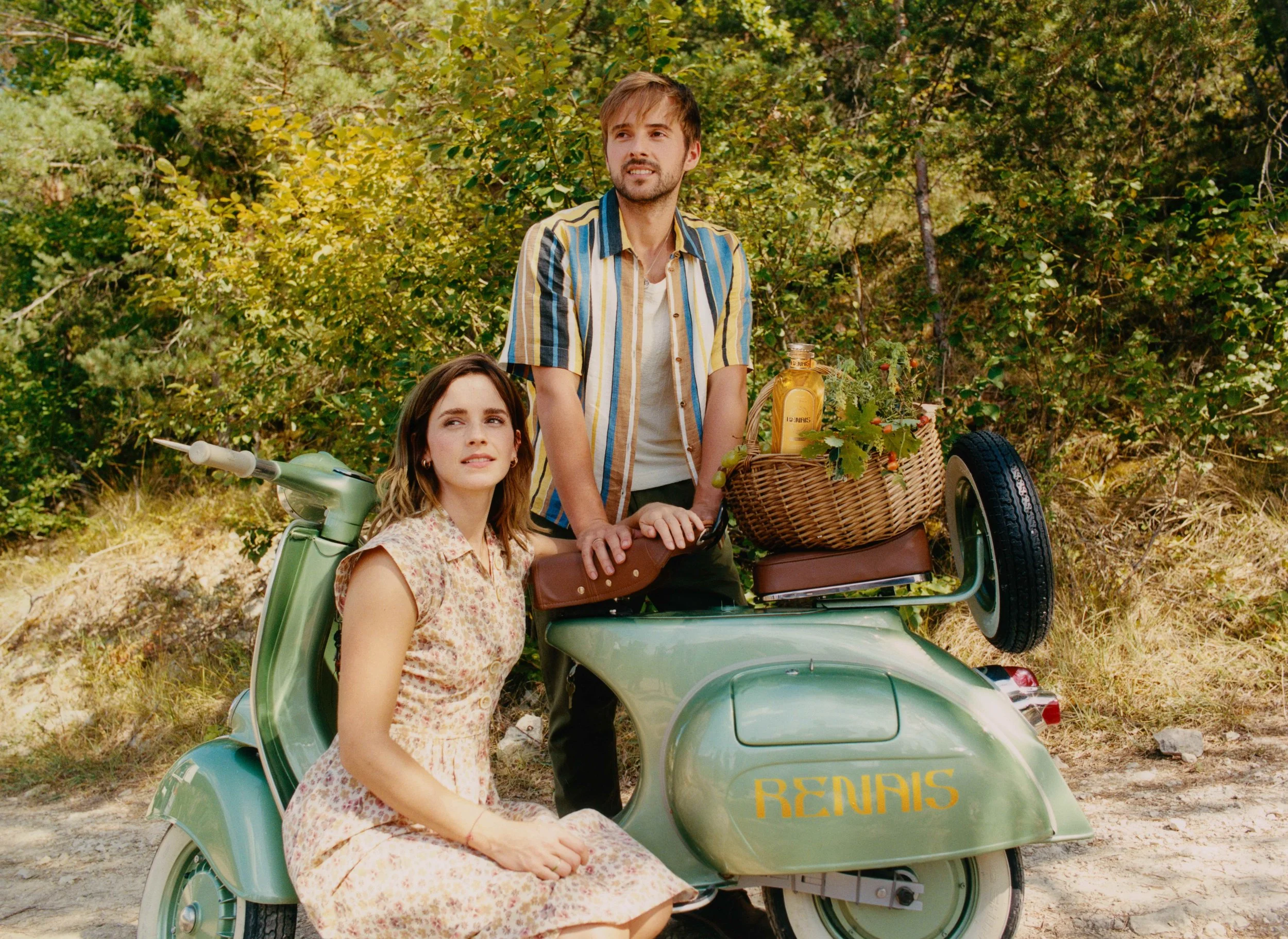 A young woman in a floral dress sitting on a vintage green scooter with a man standing next to her, both in a forested outdoor setting. The scooter has a wicker basket with a bottle of yellow liquid and some greenery on the back. The word 'RENAIS' is written in yellow on the scooter's side.